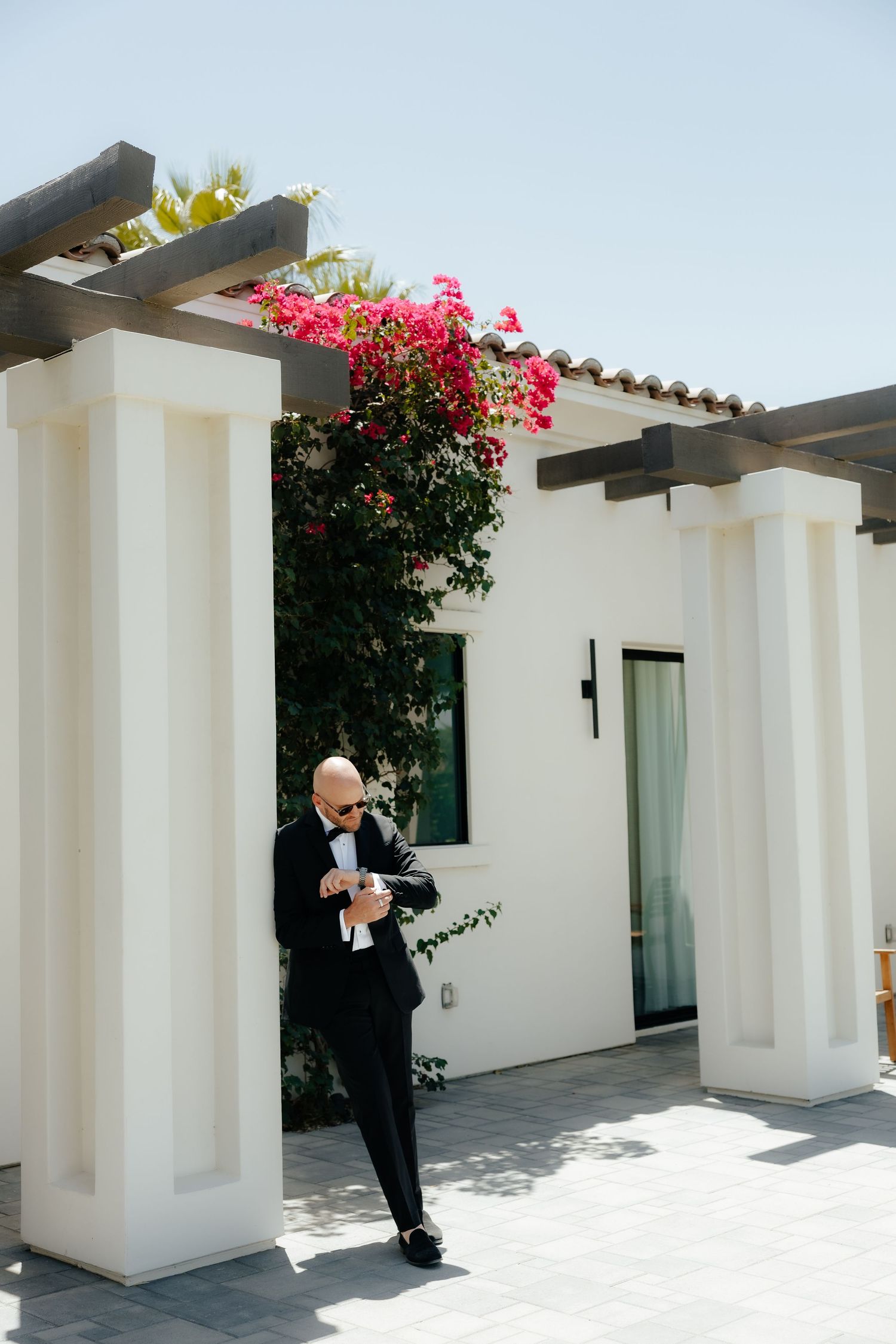 Dapper groom in black tuxedo checks his cufflinks under a white pergola adorned with vibrant pink bougainvillea.
