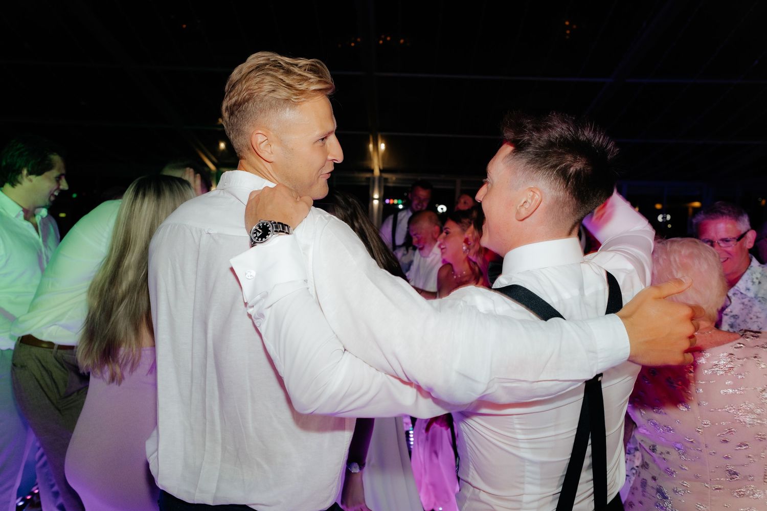 Couples dance together on a dance floor illuminated with colorful pink and purple lighting during an evening event.