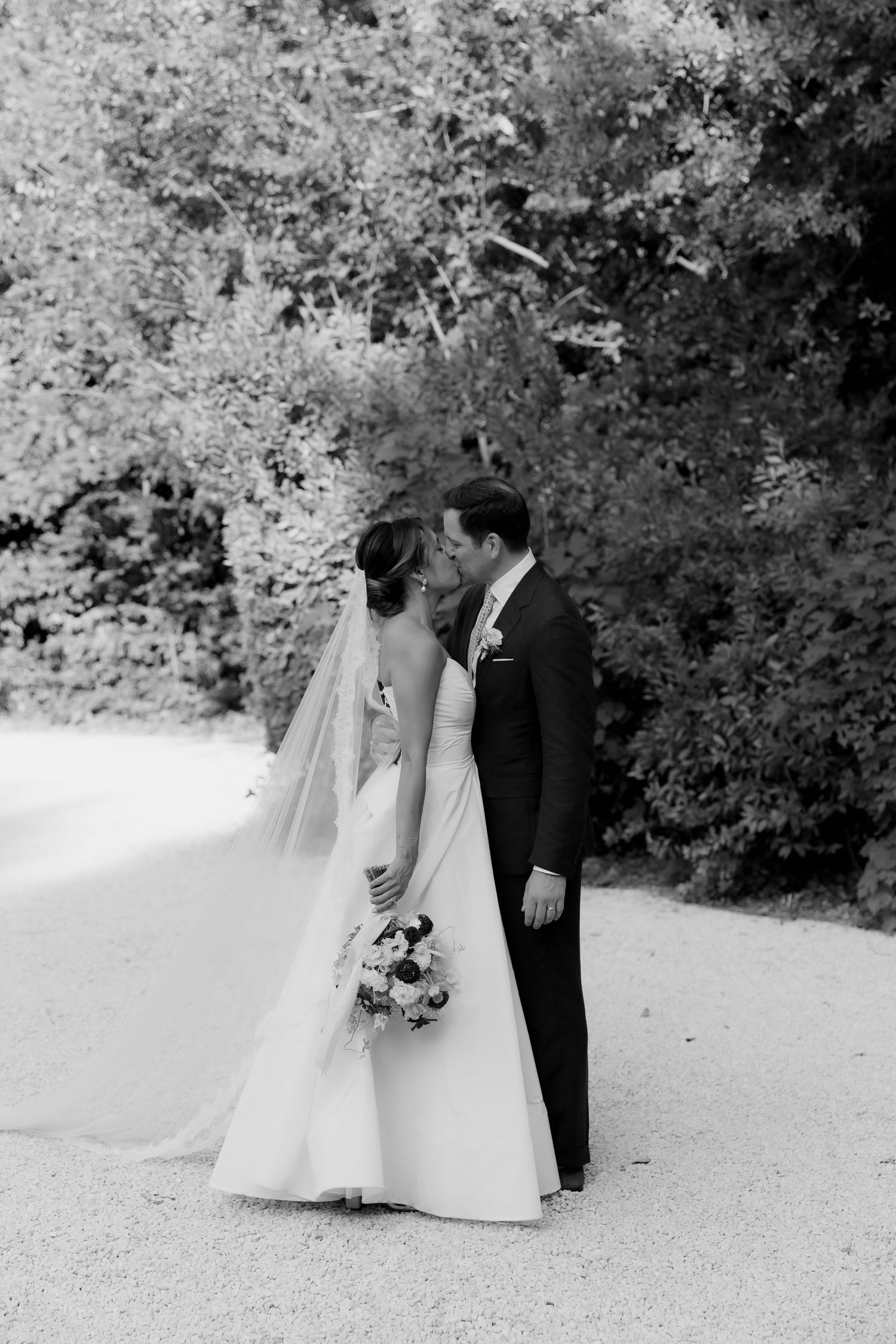 Black and white wedding portrait captured on gravel path surrounded by trees.