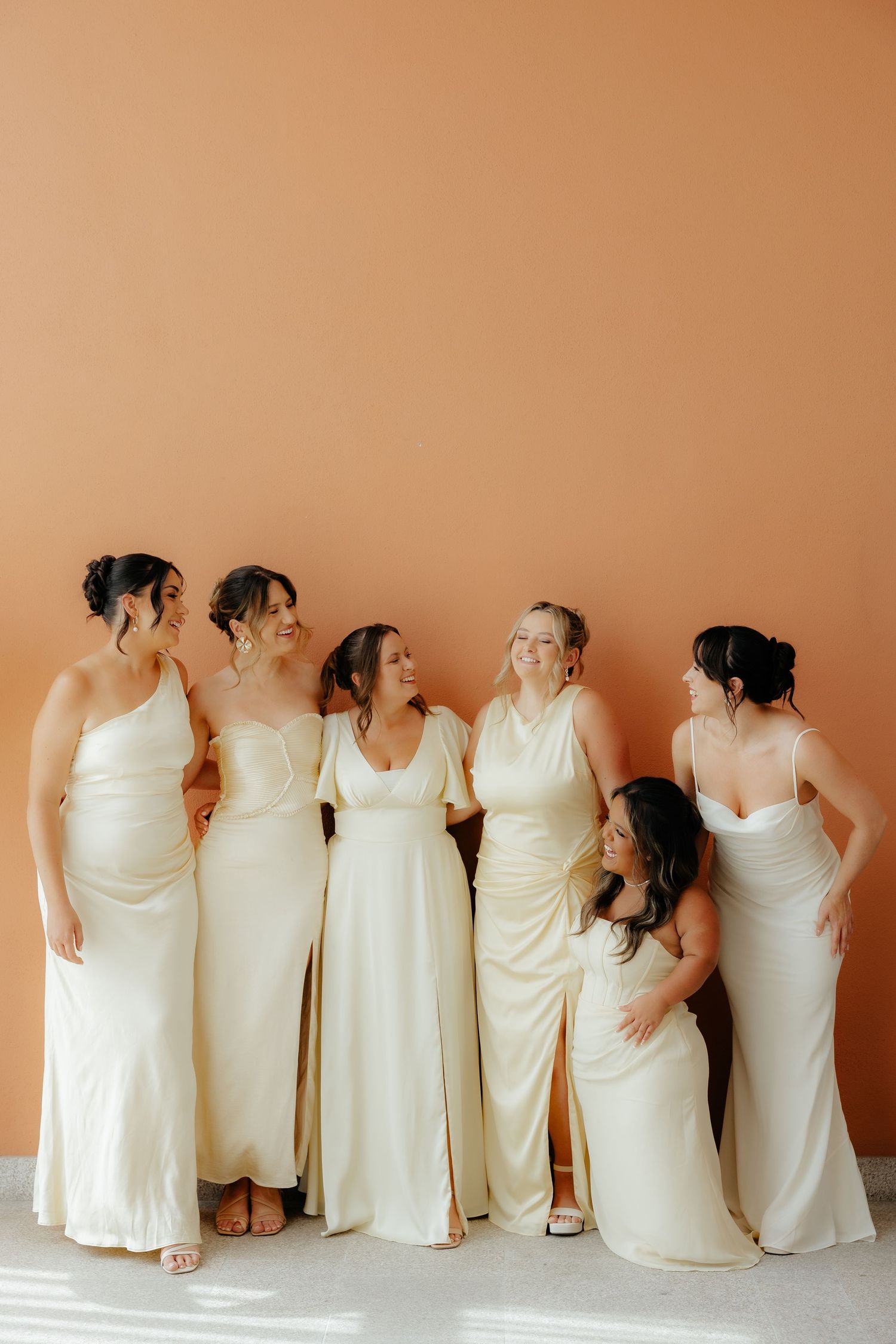 A group of bridesmaids in white dresses pose with their backs turned to the camera against a peach-colored wall.