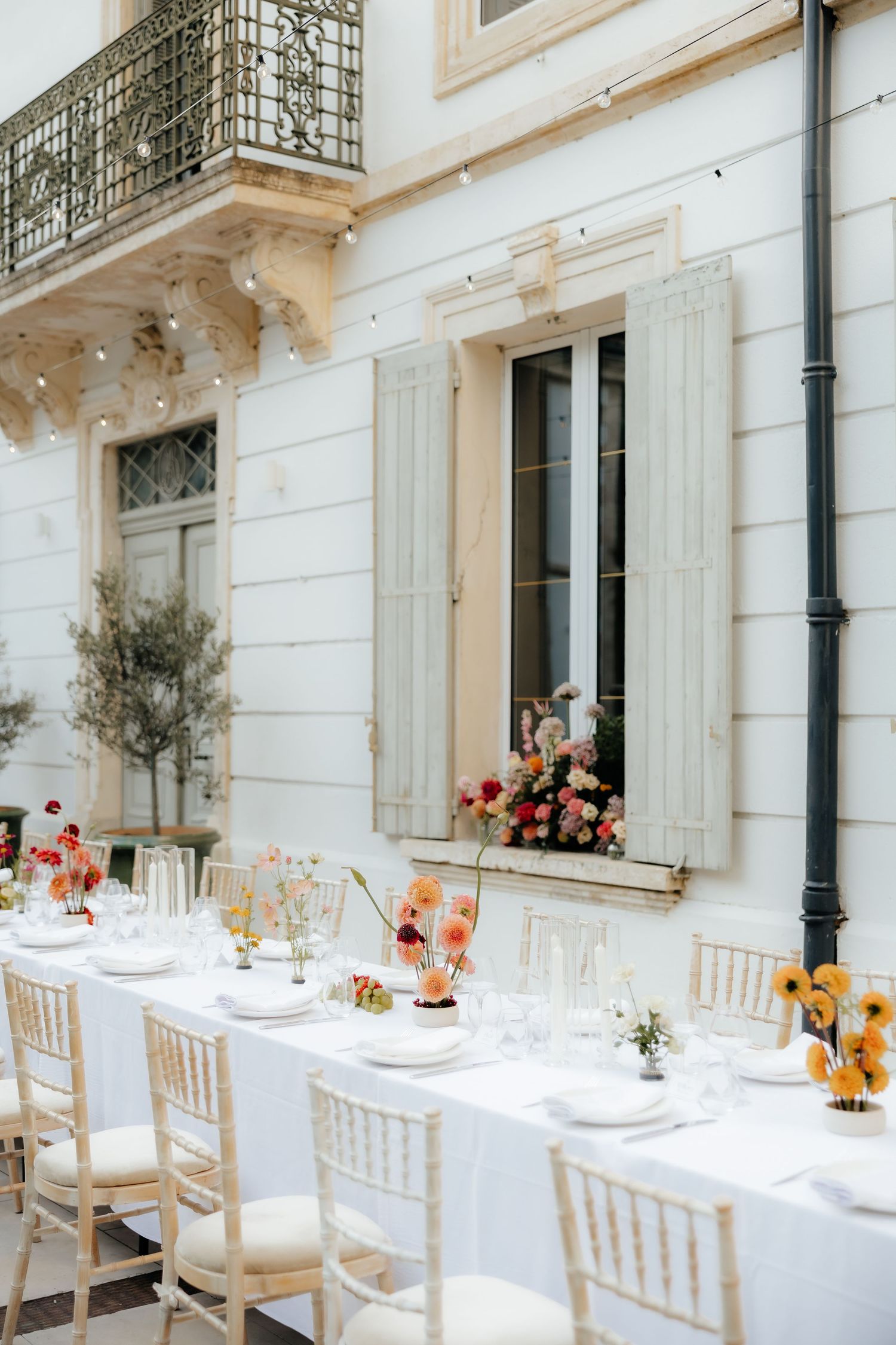 Elegant outdoor wedding reception setup with white tables and chairs along a classic building facade with floral centerpieces.