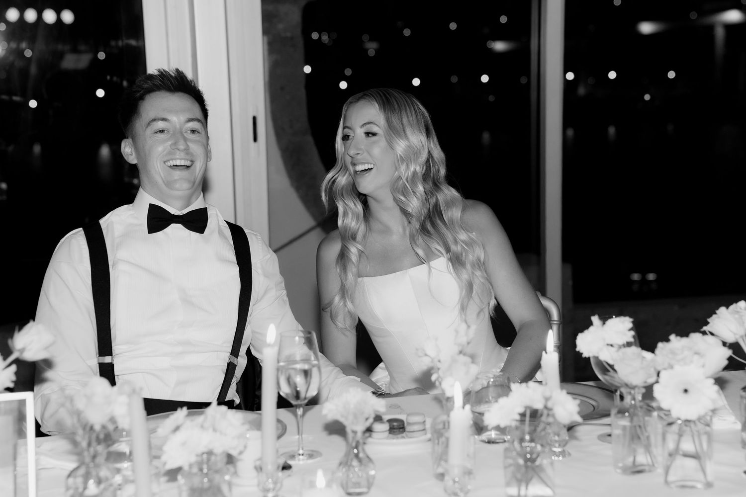 Wedding guests laugh and enjoy themselves while seated at an elegant reception dinner table with white floral centerpieces.