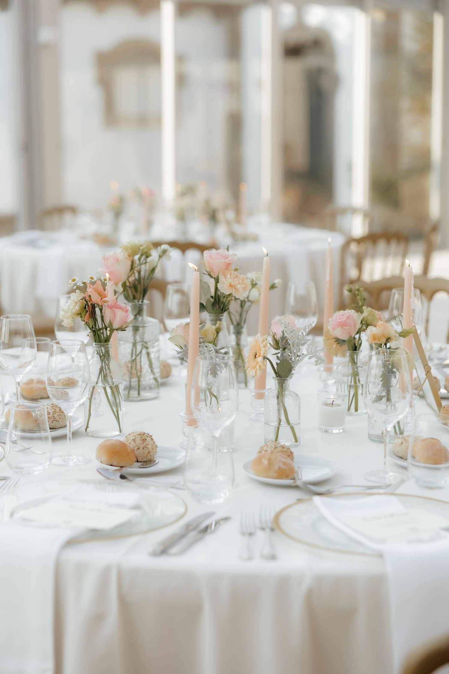 Elegant wedding reception tables set with white linens, delicate pink floral centerpieces and place settings in a bright venue.