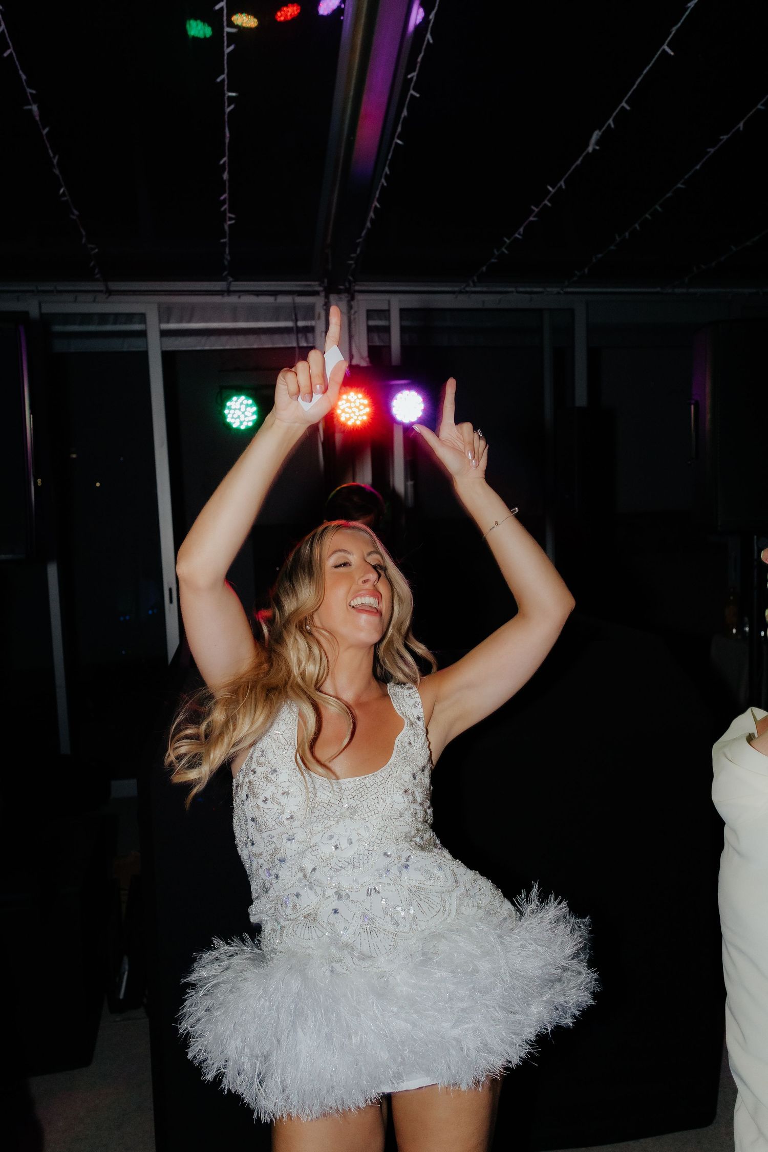 A person in a white feathered dress dances energetically at a nightclub under colorful lights.