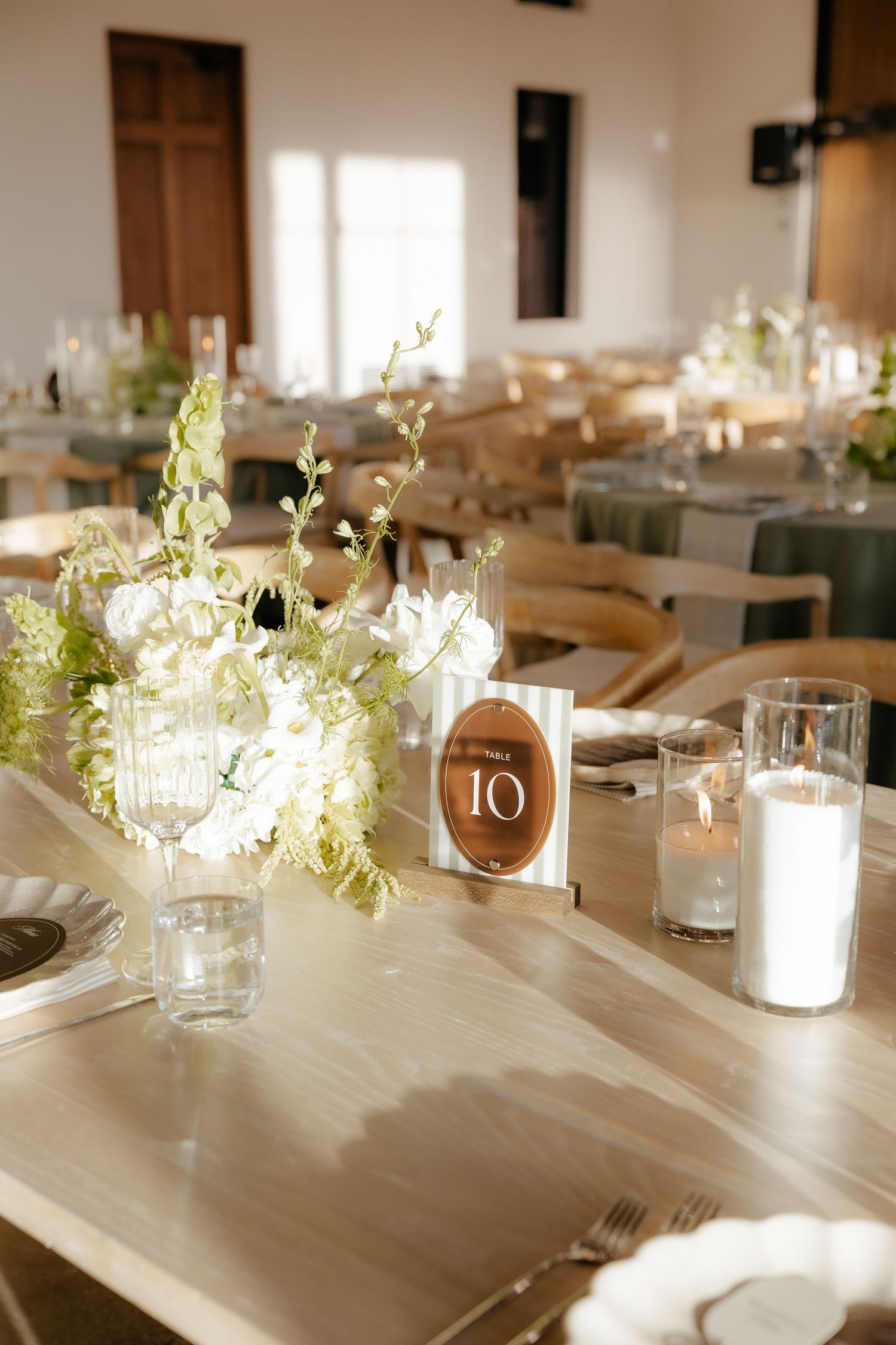 Wooden reception table centerpiece featuring white flowers, greenery, candles, and brown oval table number 10.