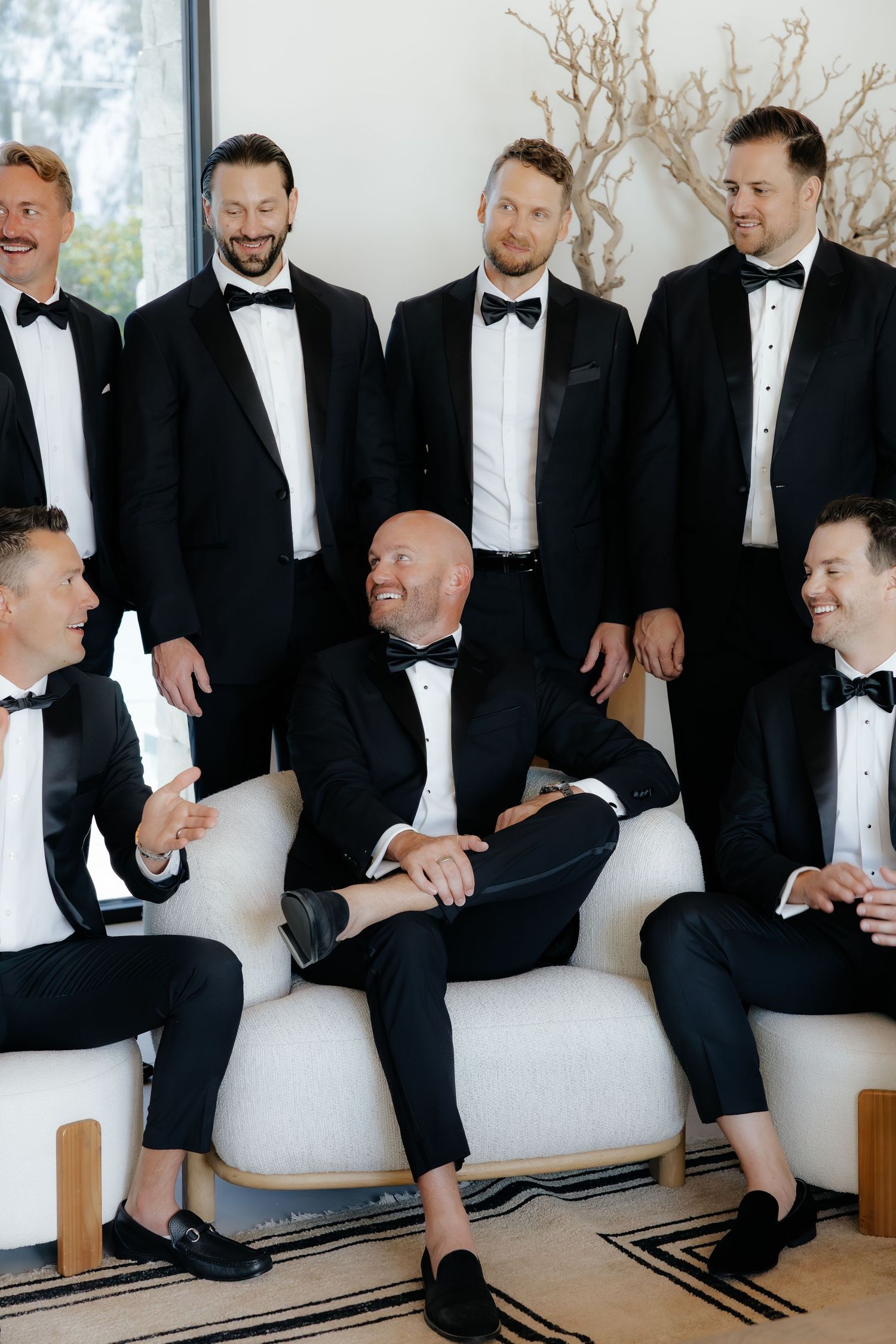 Groomsmen in black tuxedos laughing together on white sofa during wedding preparations.