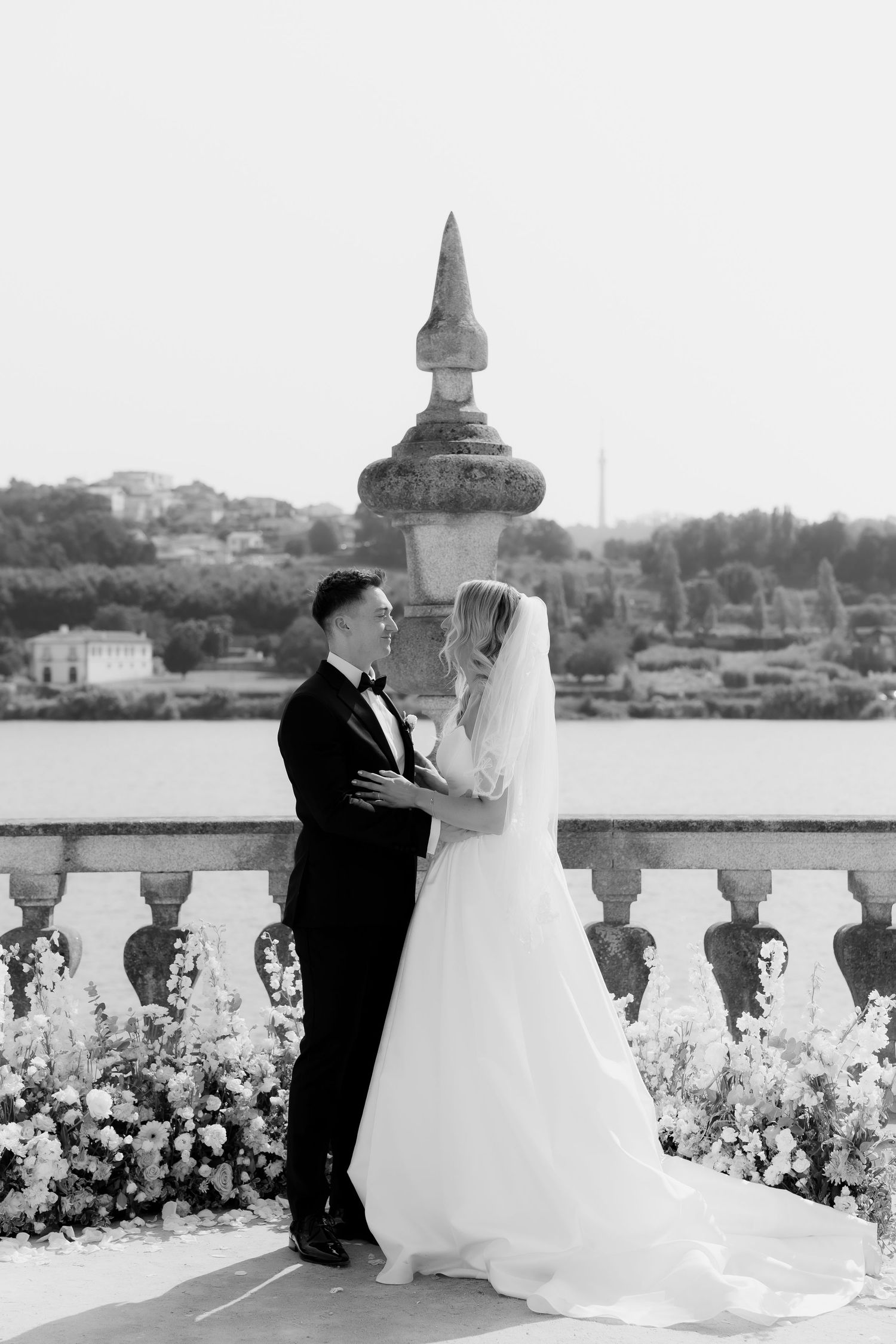 A romantic wedding couple embraces on a waterfront terrace with a scenic harbor view in the background.