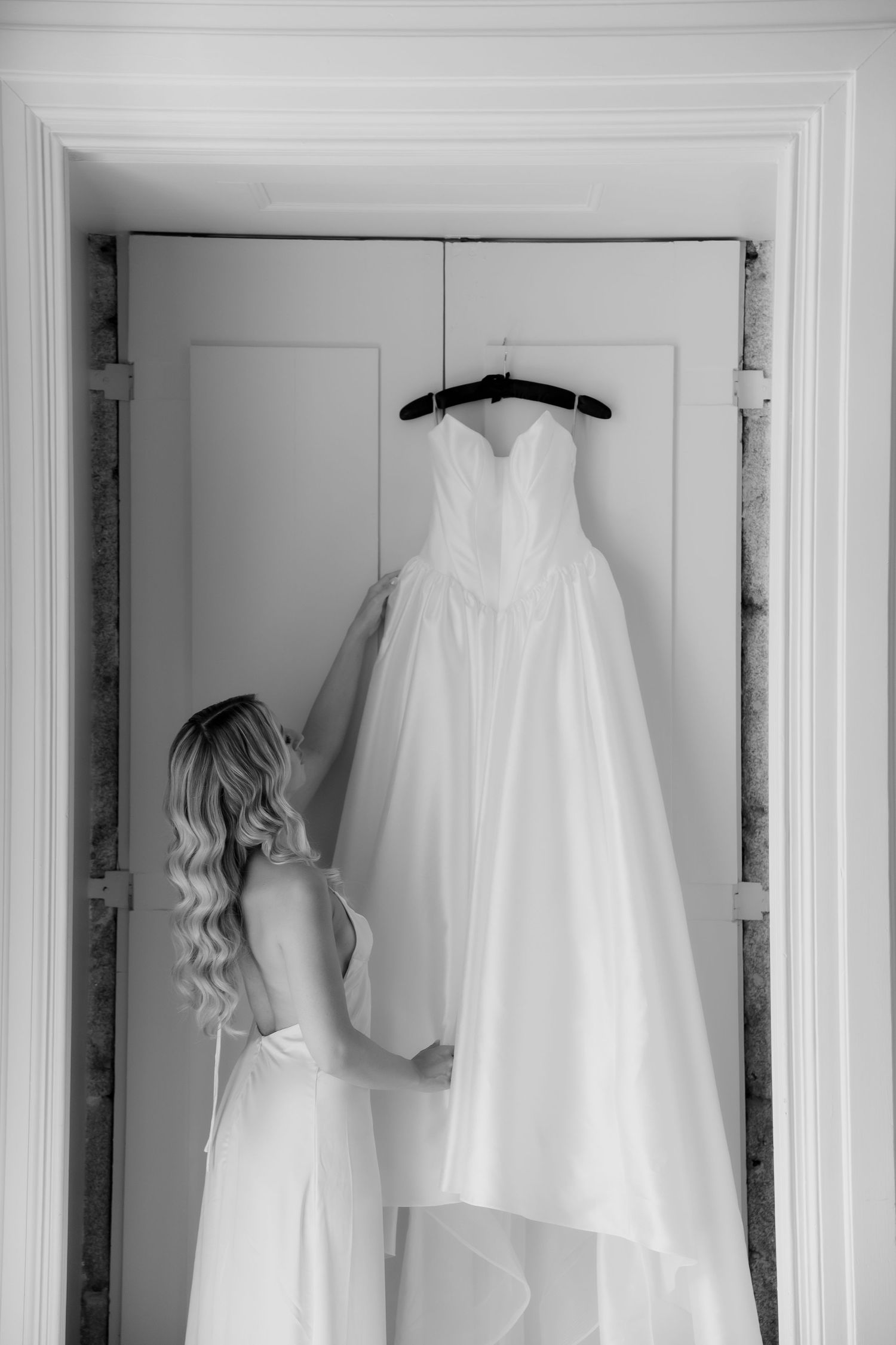 Black and white photo of bride with curly hair examining her wedding dress on hanger.