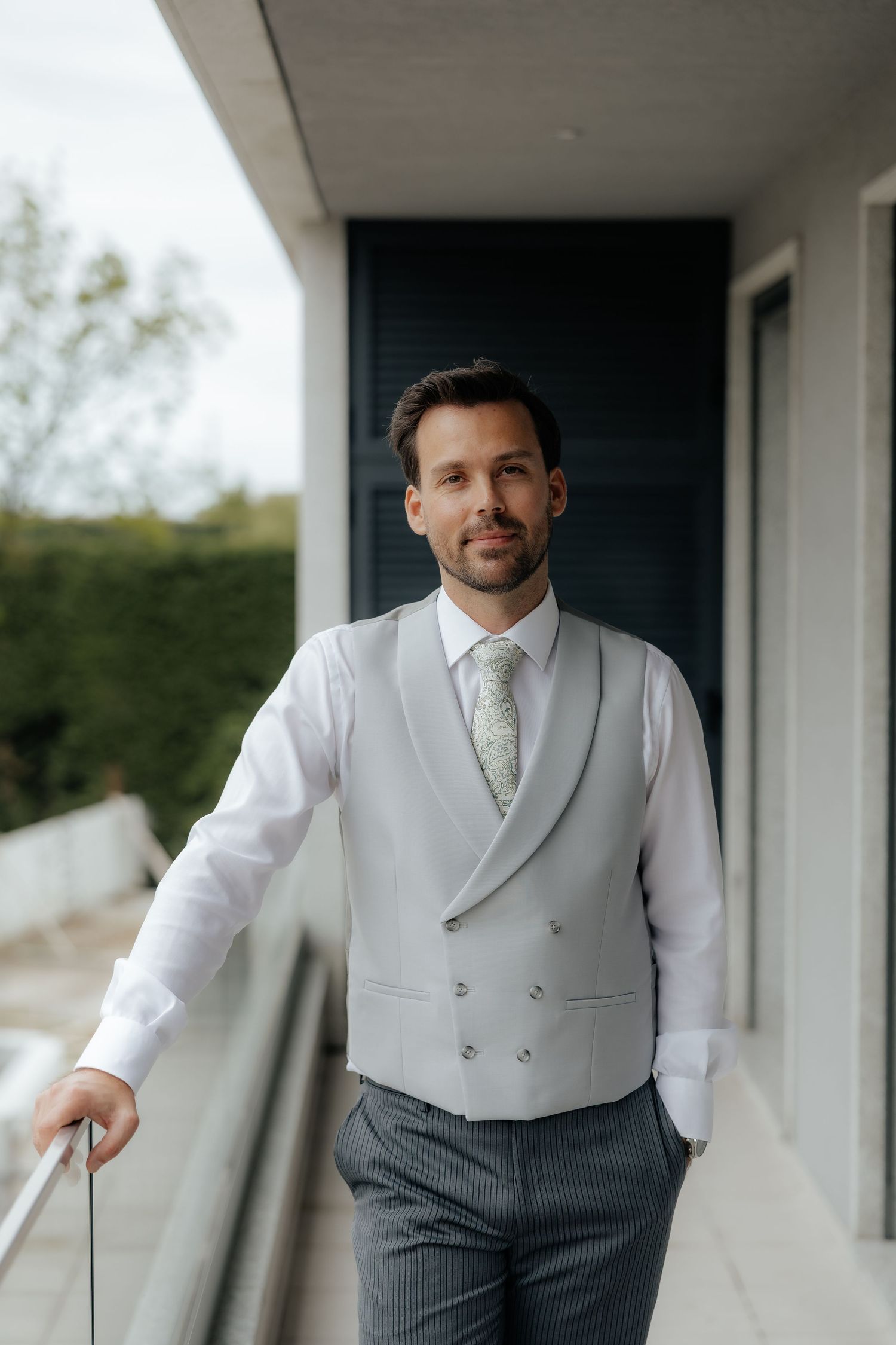 Groom in gray vest and striped trousers stands on balcony holding railing.
