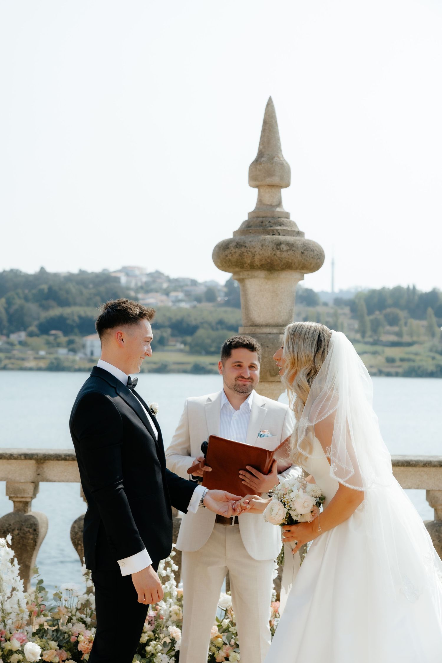 A wedding ceremony takes place on a scenic waterfront terrace with ornate stone balustrades and floral arrangements.
