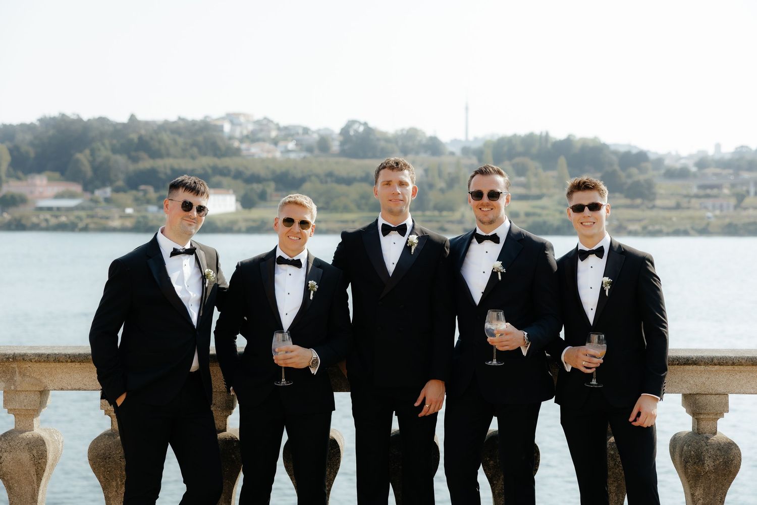 Groomsmen in black tuxedos and sunglasses pose together on a waterfront terrace with ocean views in the background.