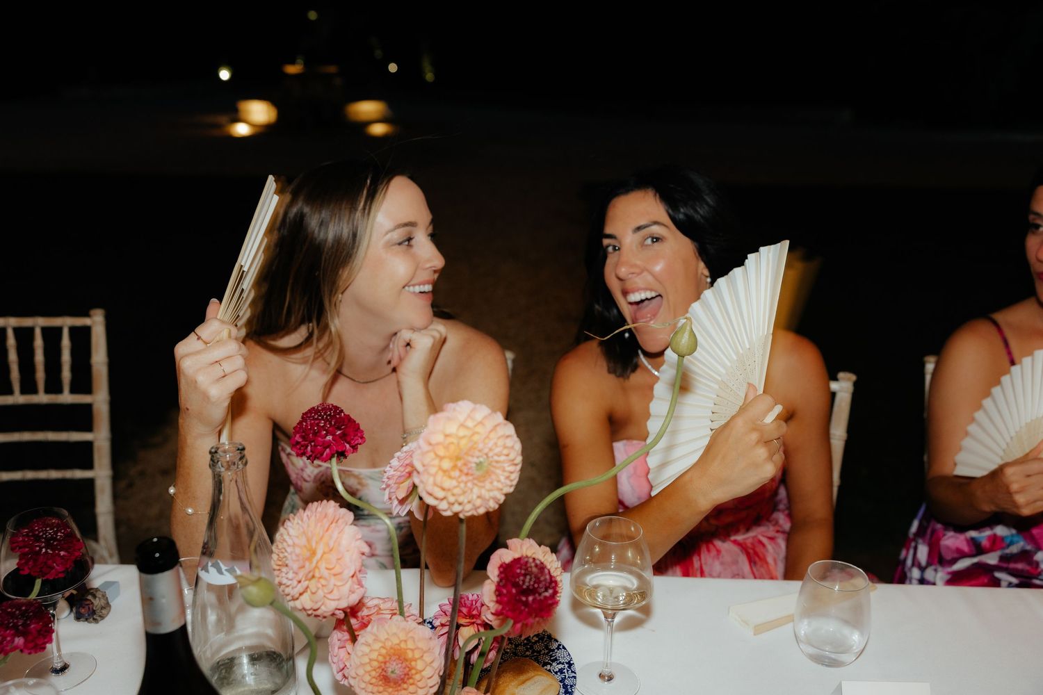Wedding guests engage in animated conversation at a reception dinner table decorated with pink floral centerpieces.