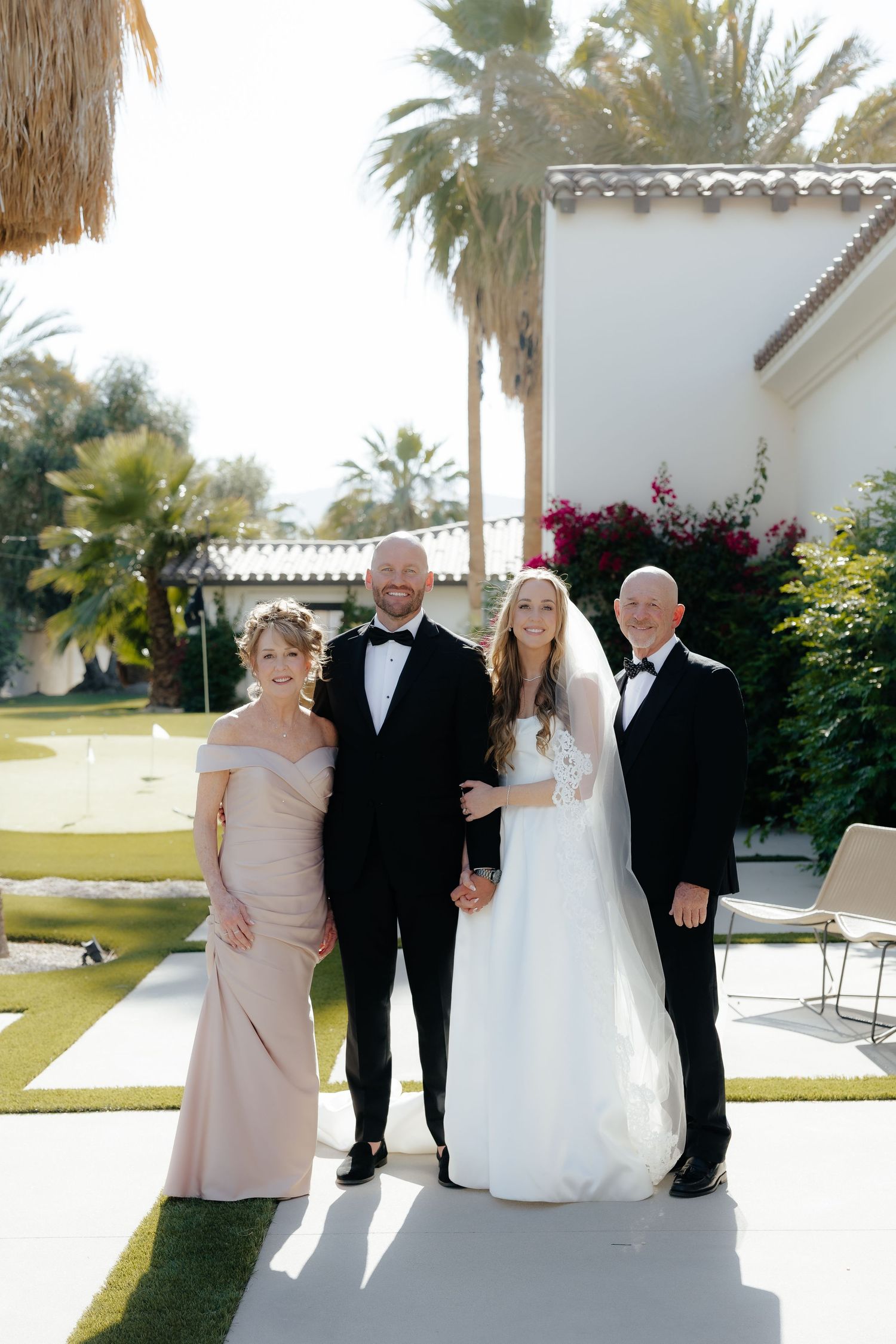 Wedding party group photo with bride, groom, and two guests in formal attire outdoors.