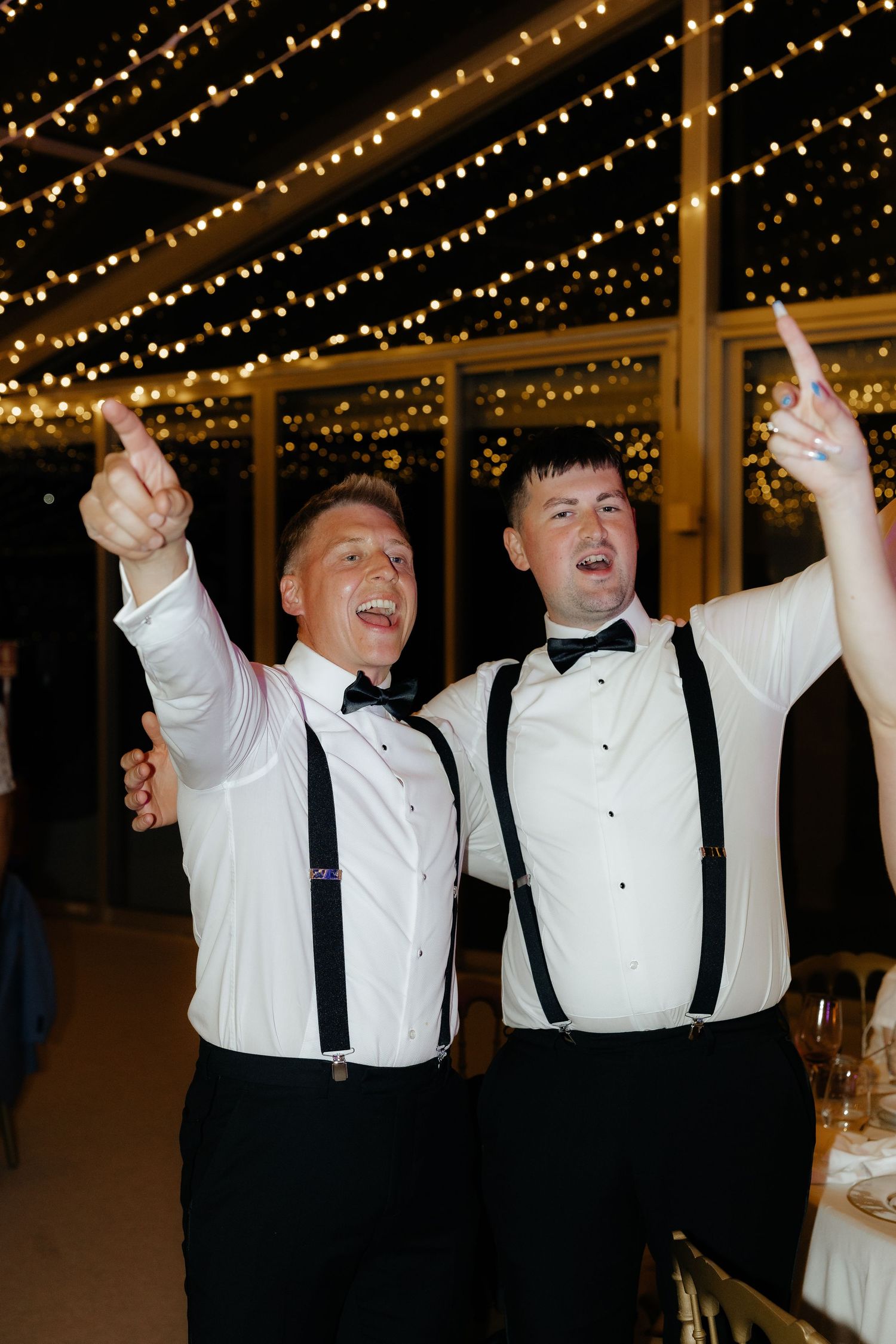 Wedding party guests in black and white formal attire celebrating joyfully with raised arms under twinkling lights.