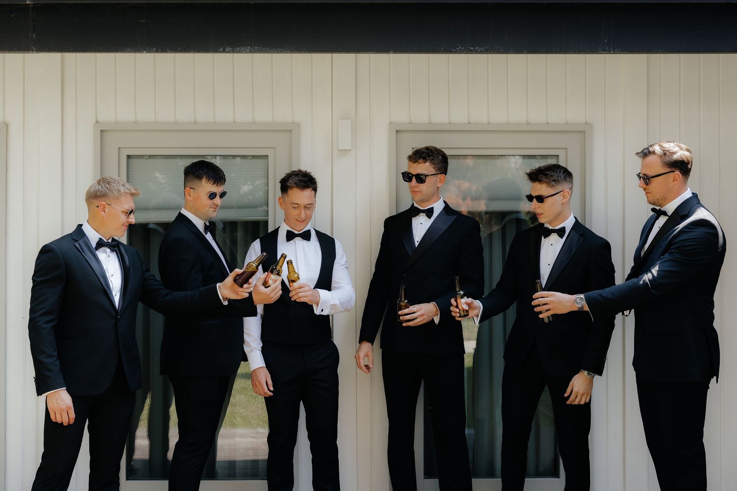 Groomsmen in black tuxedos and sunglasses gather for drinks before wedding ceremony.