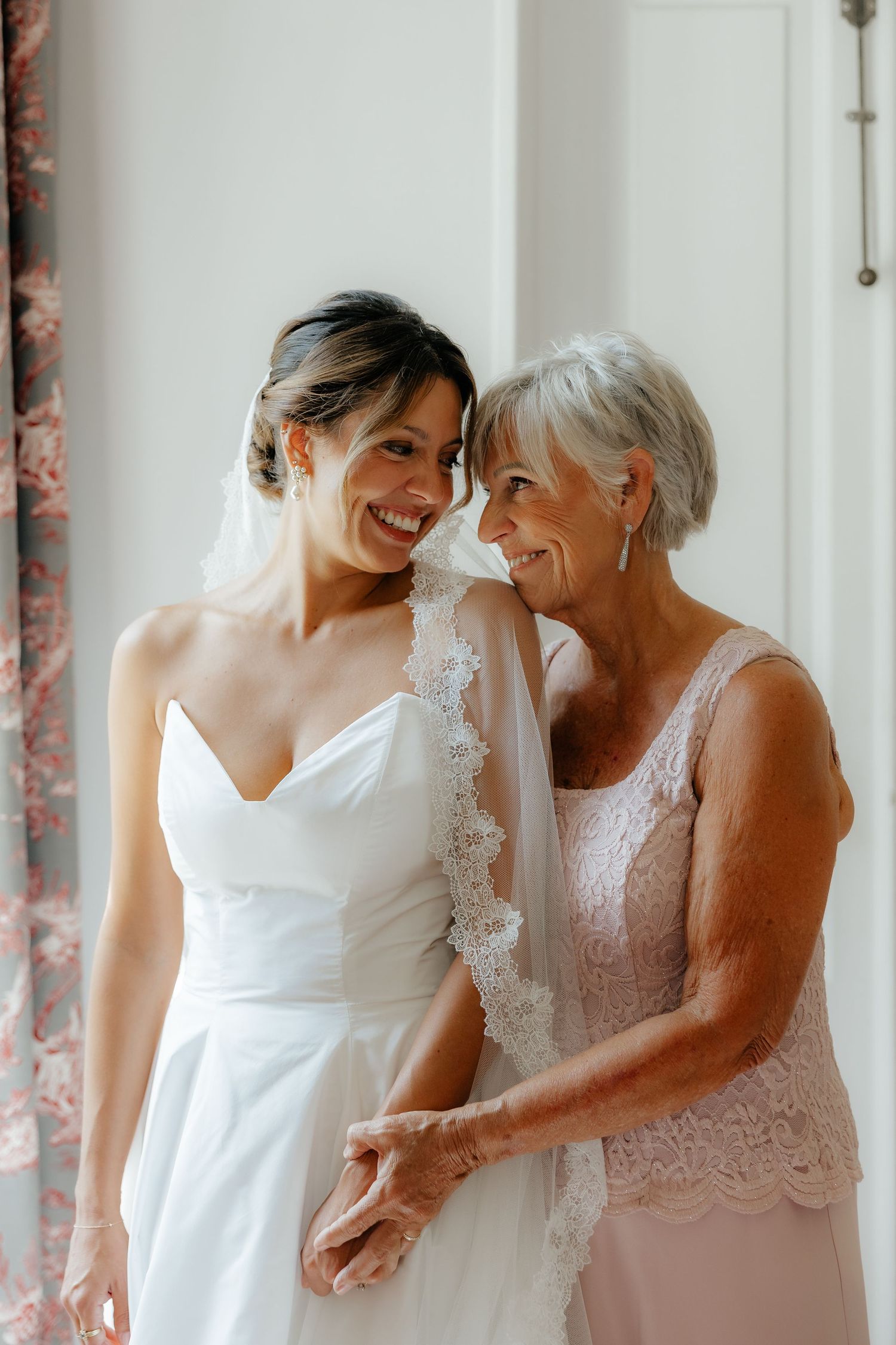 Intimate wedding day moment shared between bride in white dress and woman in pink lace dress.