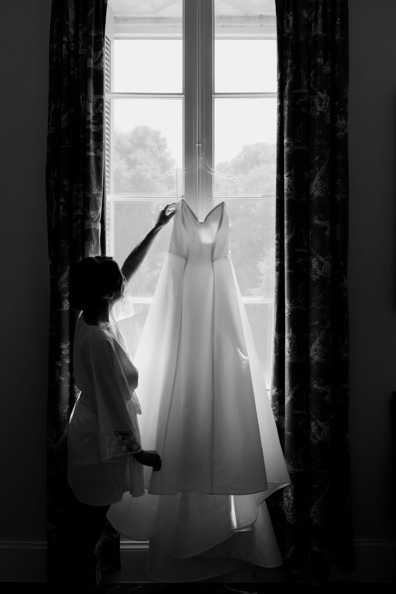 Silhouette of wedding dress preparation against window with dark curtains.