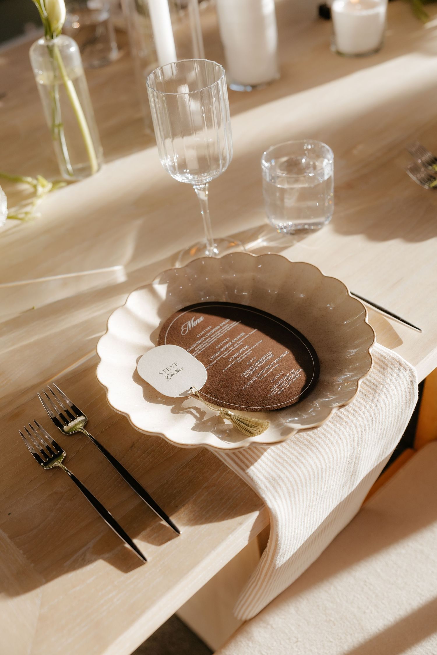 Elegant wedding table setting with scalloped white plate, brown charger, gold cutlery and glassware.