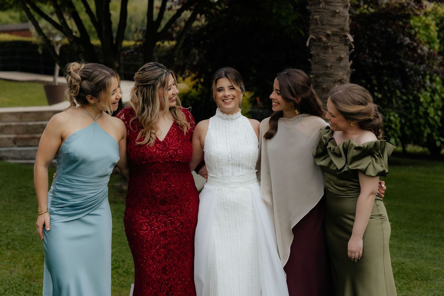 Bride smiles with four bridesmaids in colorful gowns on green lawn with palm trees behind.