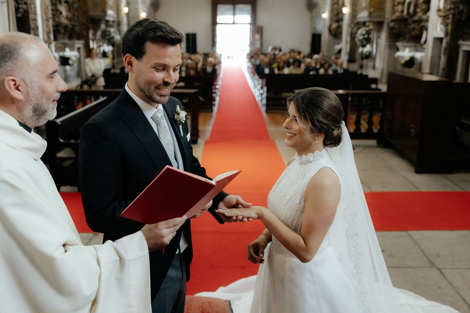 Priest officiates wedding ceremony as groom places ring on brides finger at church altar.