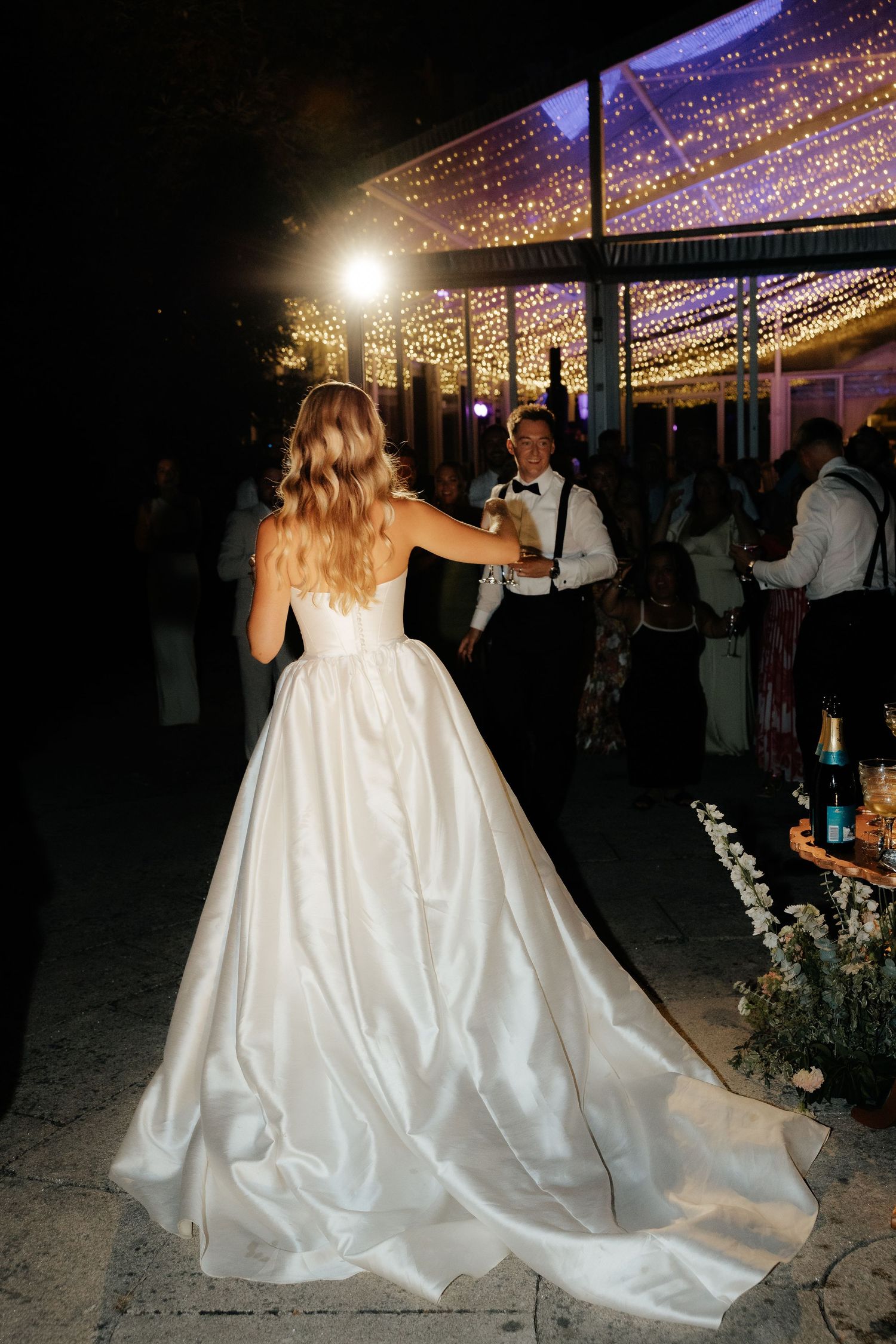 Bride in elegant ballgown with long train walks toward groom under twinkling lights at reception.