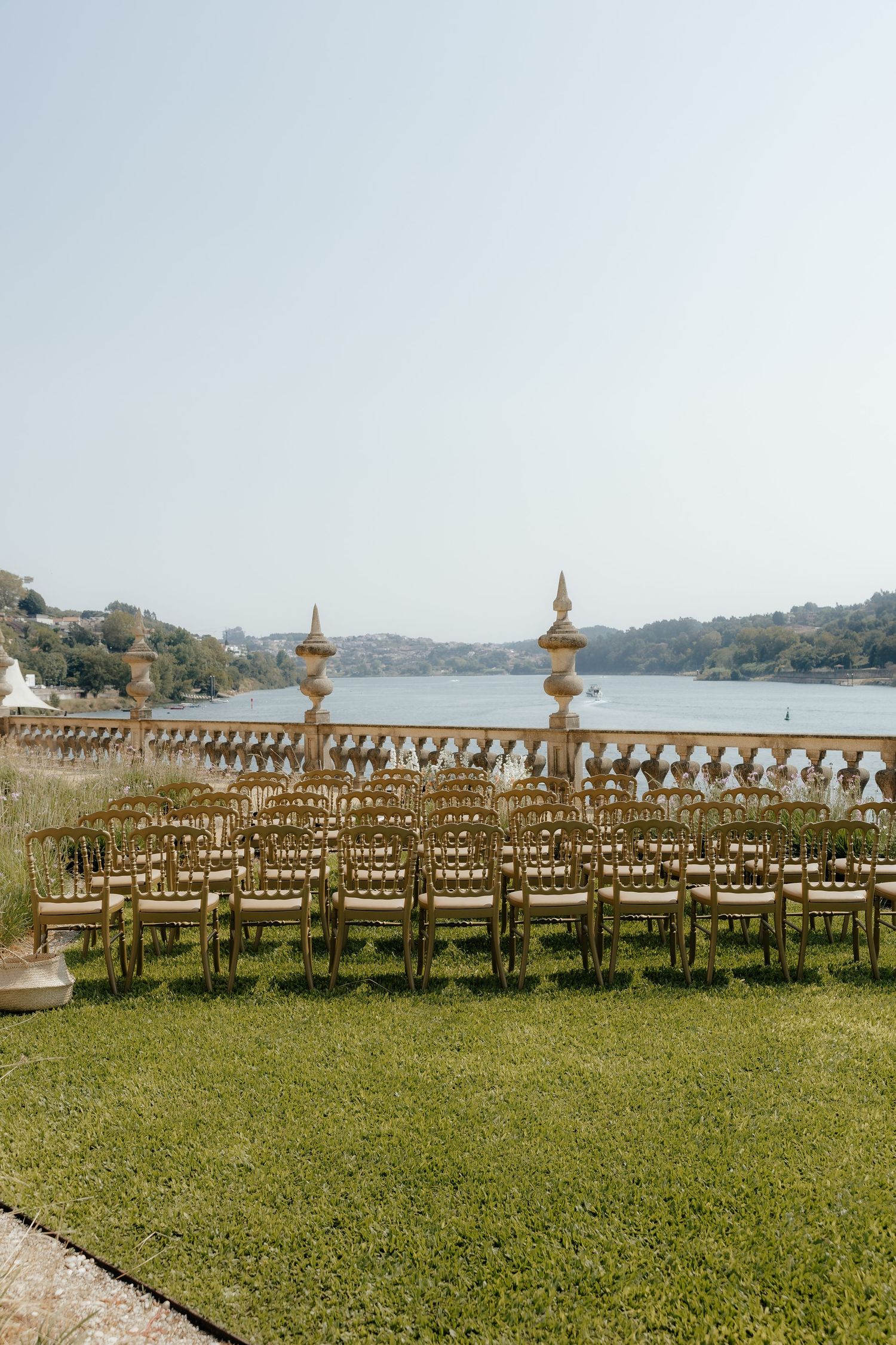 Rows of gold Chiavari chairs set on green lawn overlooking a river for an outdoor wedding ceremony.