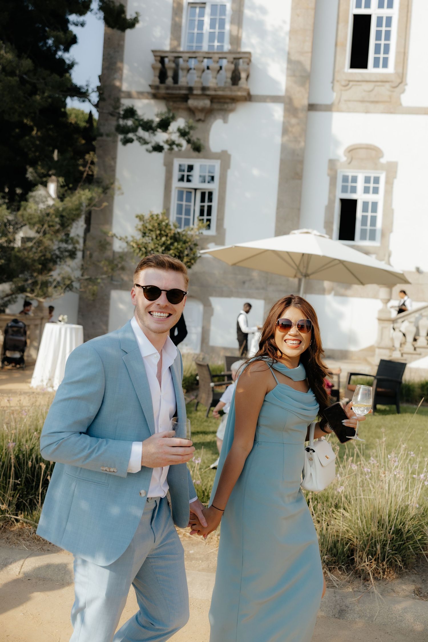 Stylish wedding guests in matching light blue outfits holding drinks and walking hand in hand outdoors.