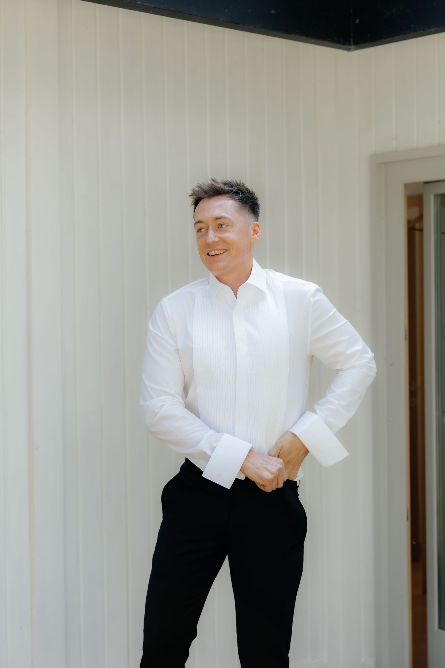 Smiling groom in a white dress shirt and black trousers standing against a white paneled wall before getting dressed.