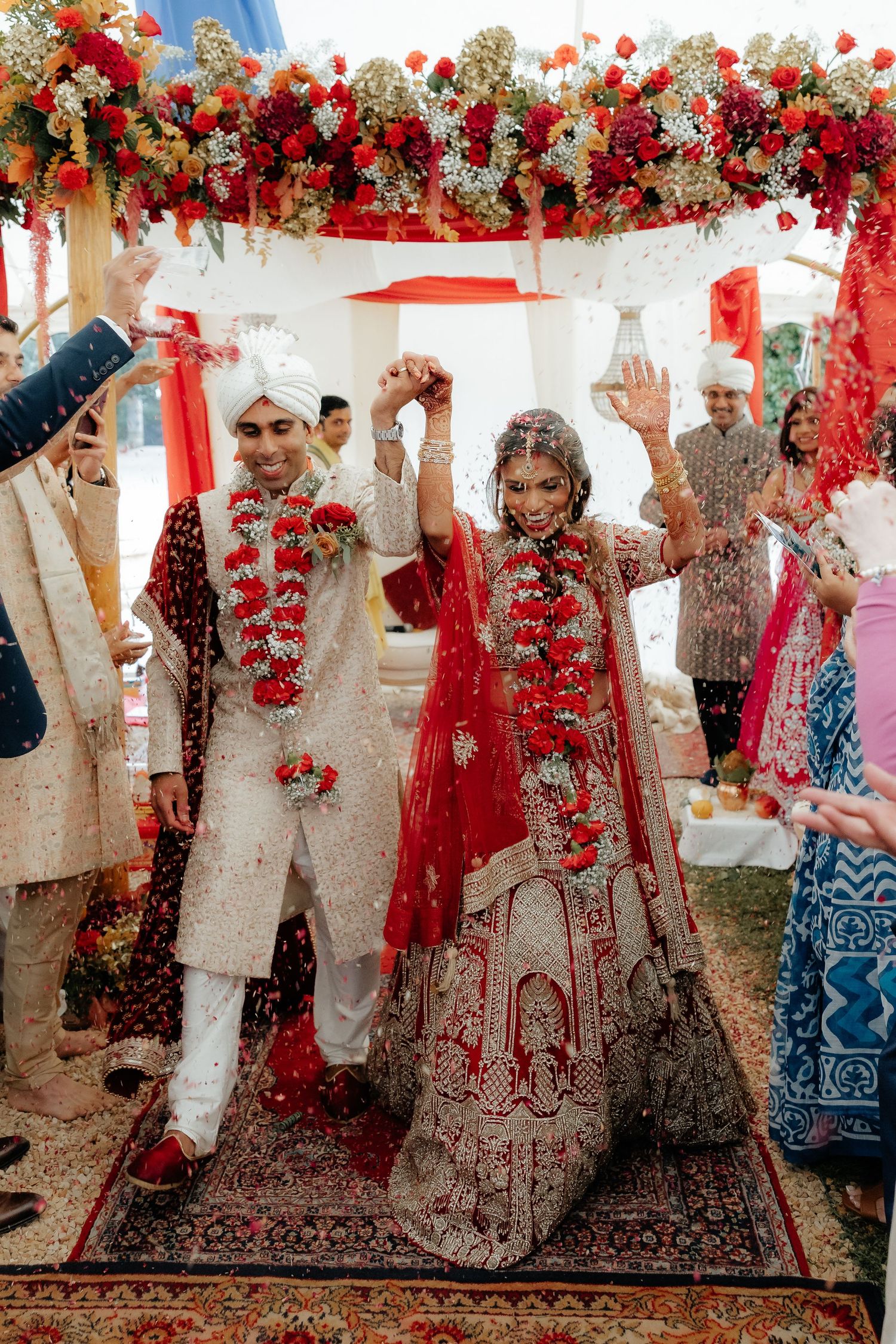 Indian bride and groom celebrate under floral mandap in red and white traditional wedding attire.
