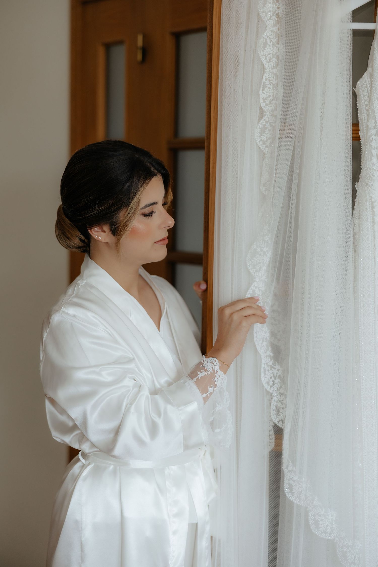 Bride in white silk robe gently touches the delicate lace trim of her hanging wedding veil.