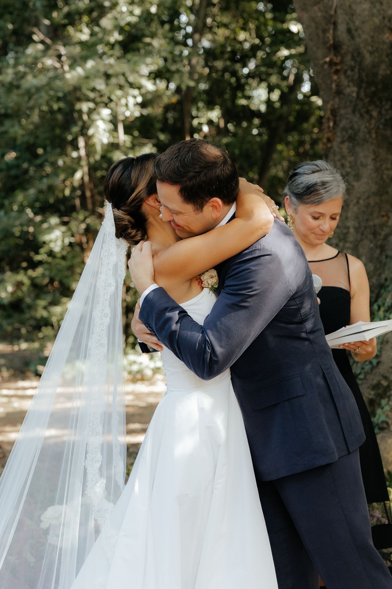 A romantic wedding moment captured under trees with flowing white veil and navy suit.
