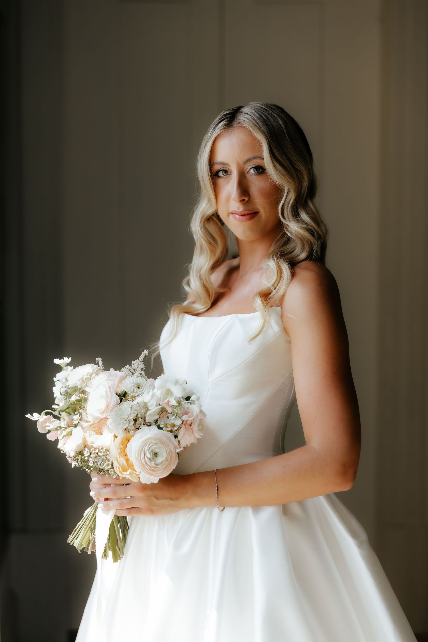 A bride in a classic white wedding dress holds a delicate bouquet of white flowers against a moody dark background.