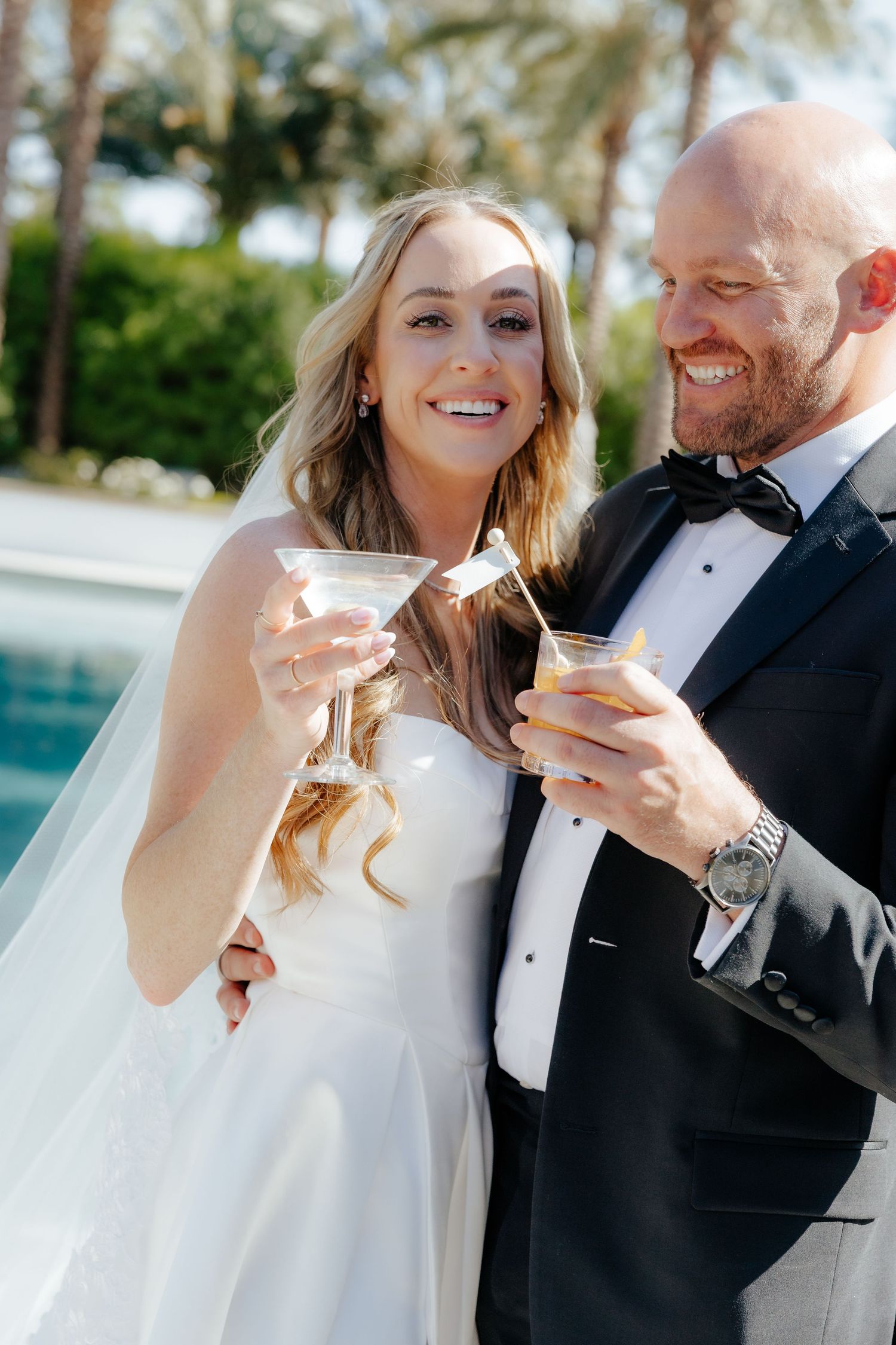 Smiling bride and groom holding cocktails near a pool at their outdoor wedding reception.