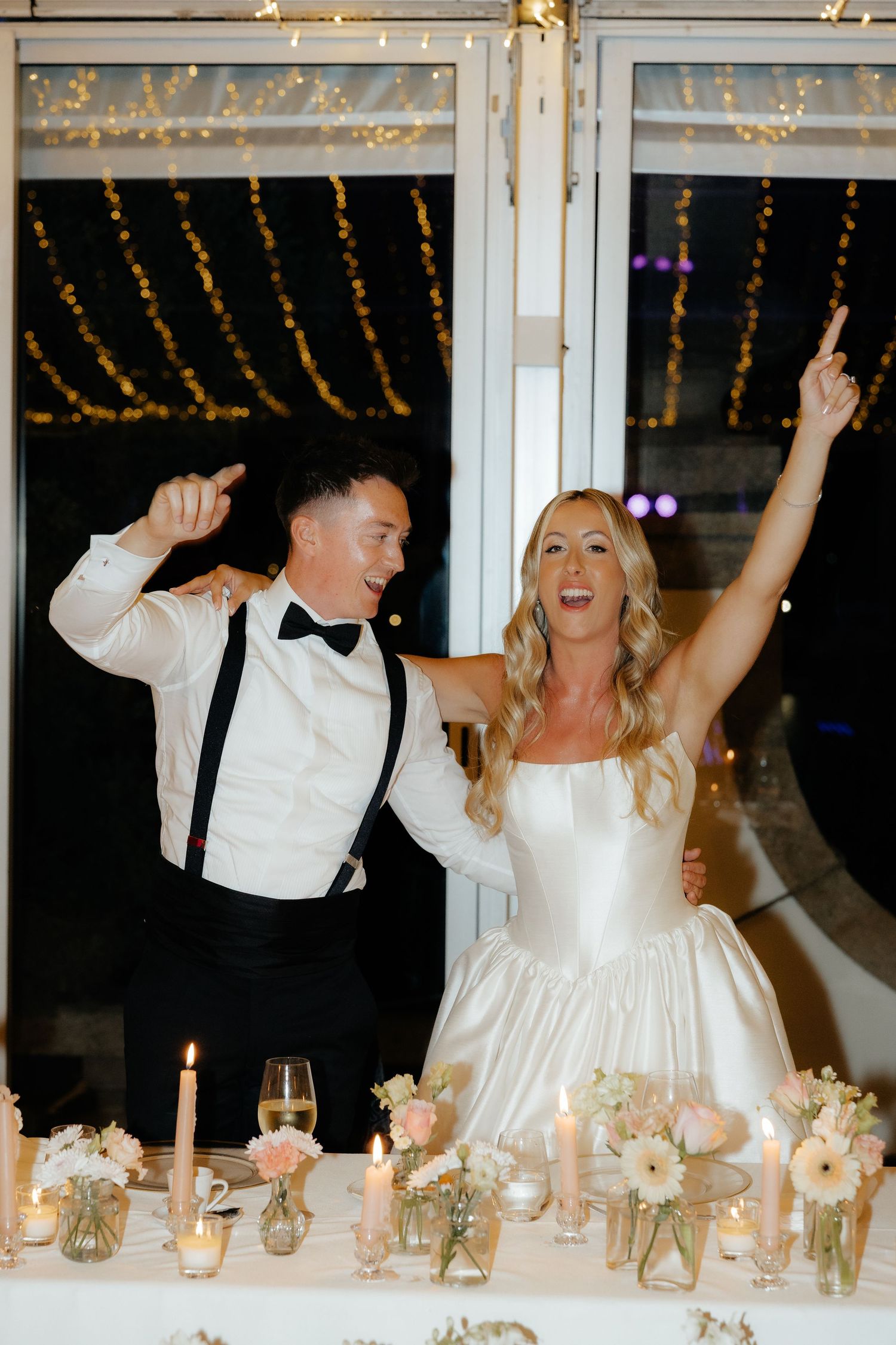 A joyful wedding celebration at a reception table with candlelight and string lights creating a festive atmosphere.