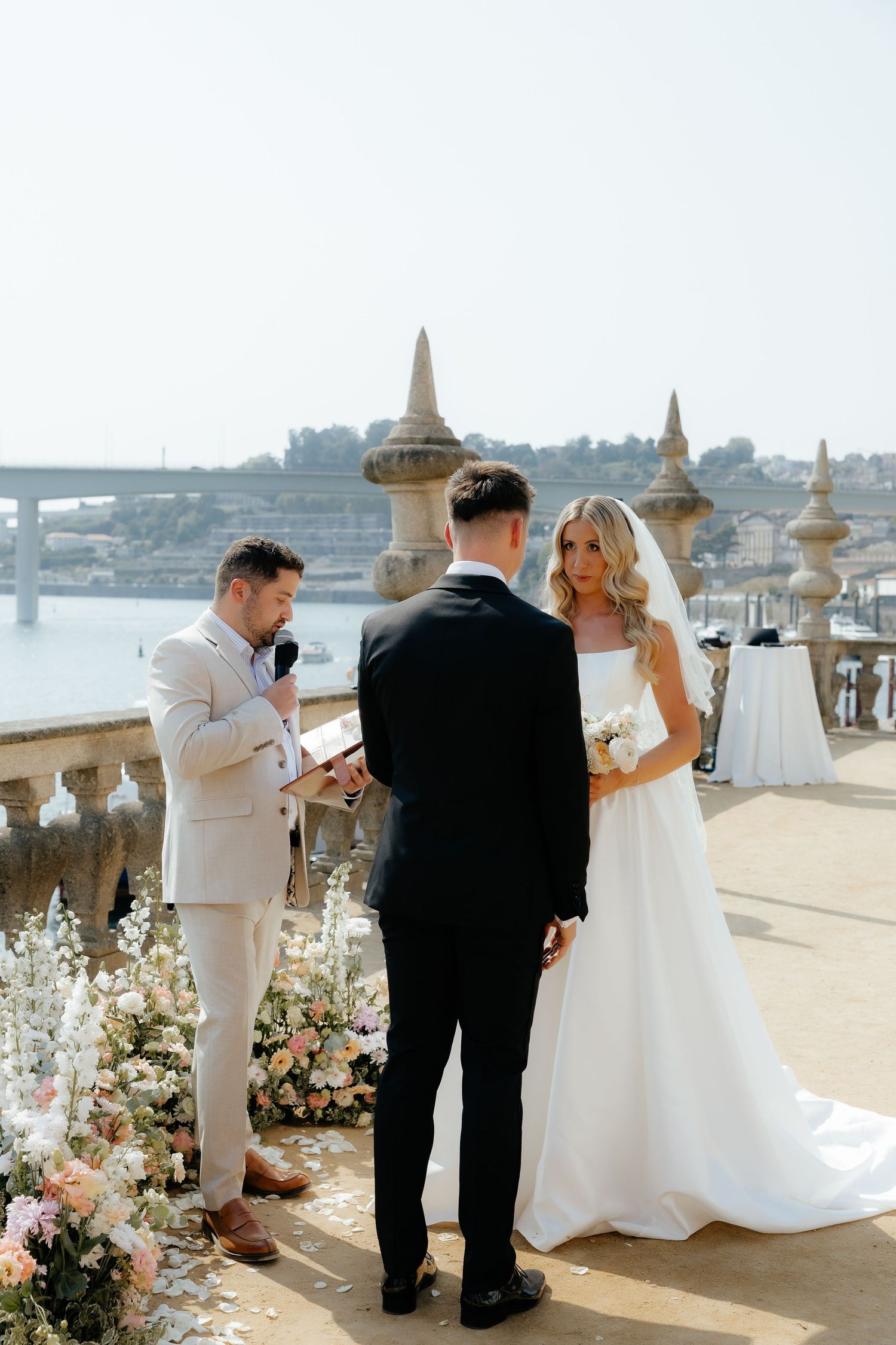 A wedding ceremony takes place on a scenic waterfront terrace decorated with white floral arrangements.