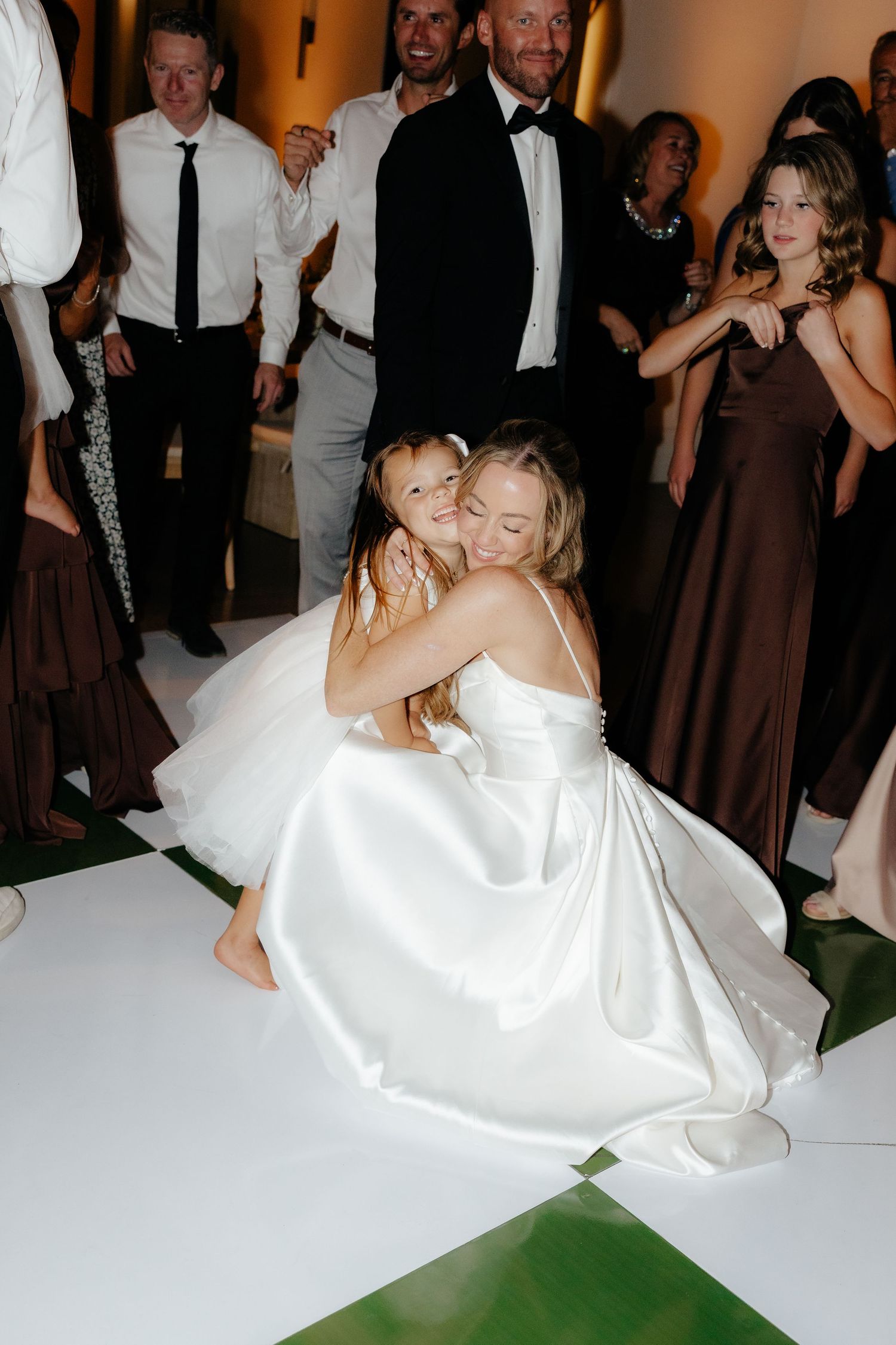 Bride in white satin gown crouches down to warmly embrace a young child on the wedding reception dance floor.