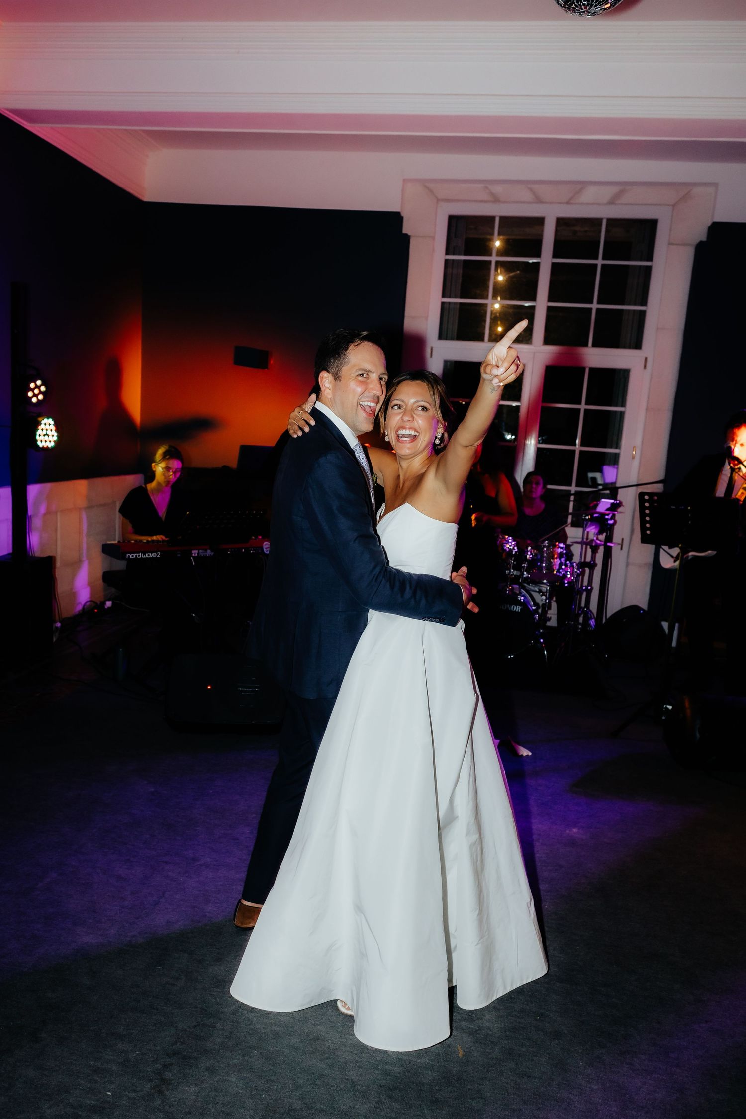 A couple in wedding attire dances together at a dimly lit reception venue with purple lighting and white decor.