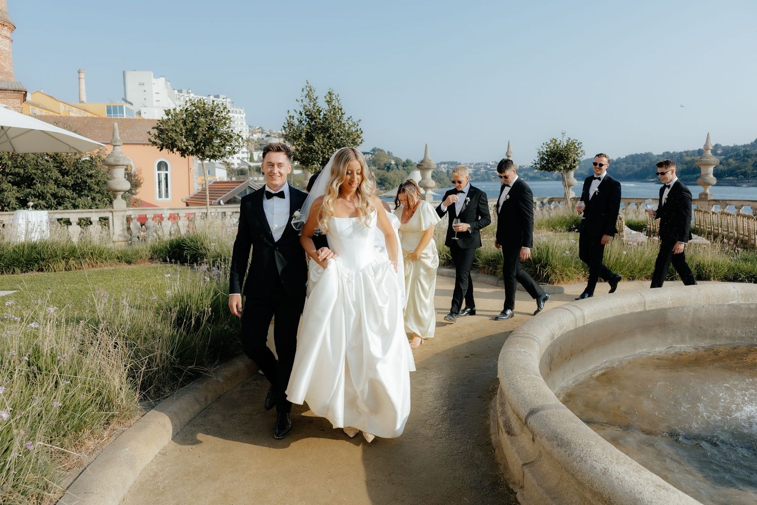 Bride in white gown and groom in tuxedo walking with wedding party near scenic waterfront fountain.