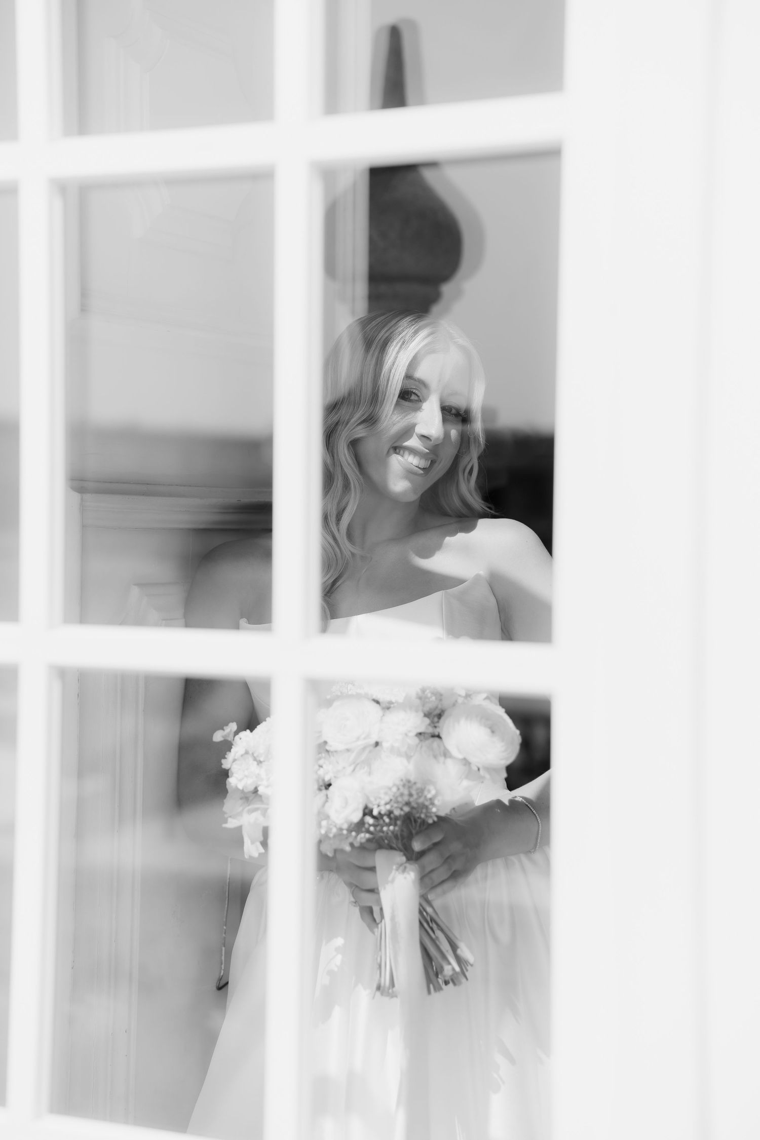 Black and white photo of smiling bride holding bouquet viewed through window panes.