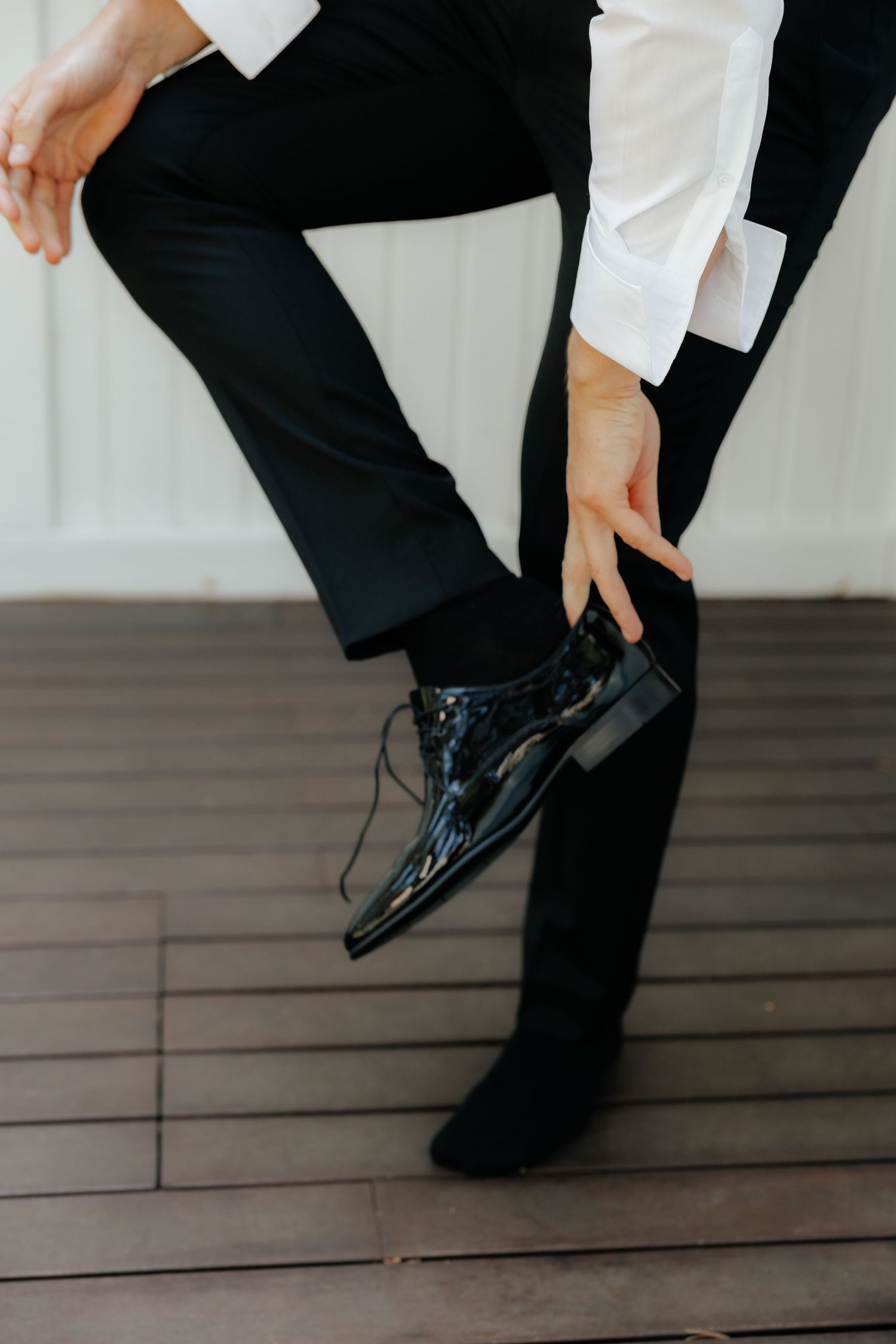 Close-up of a groom putting on shiny black patent leather dress shoes on a wooden deck floor.