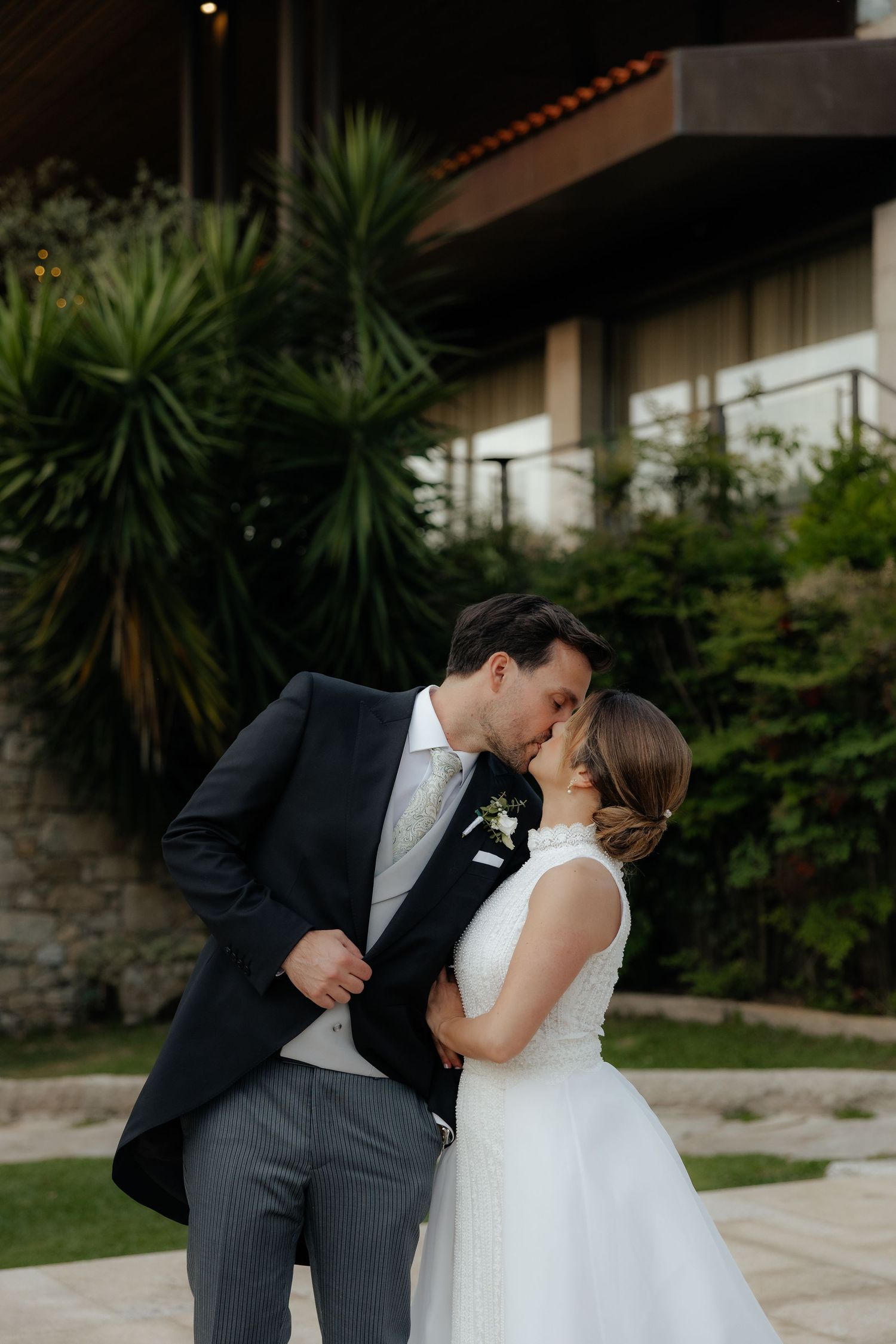 Groom in black suit leans in to kiss bride in white sleeveless gown in garden setting.