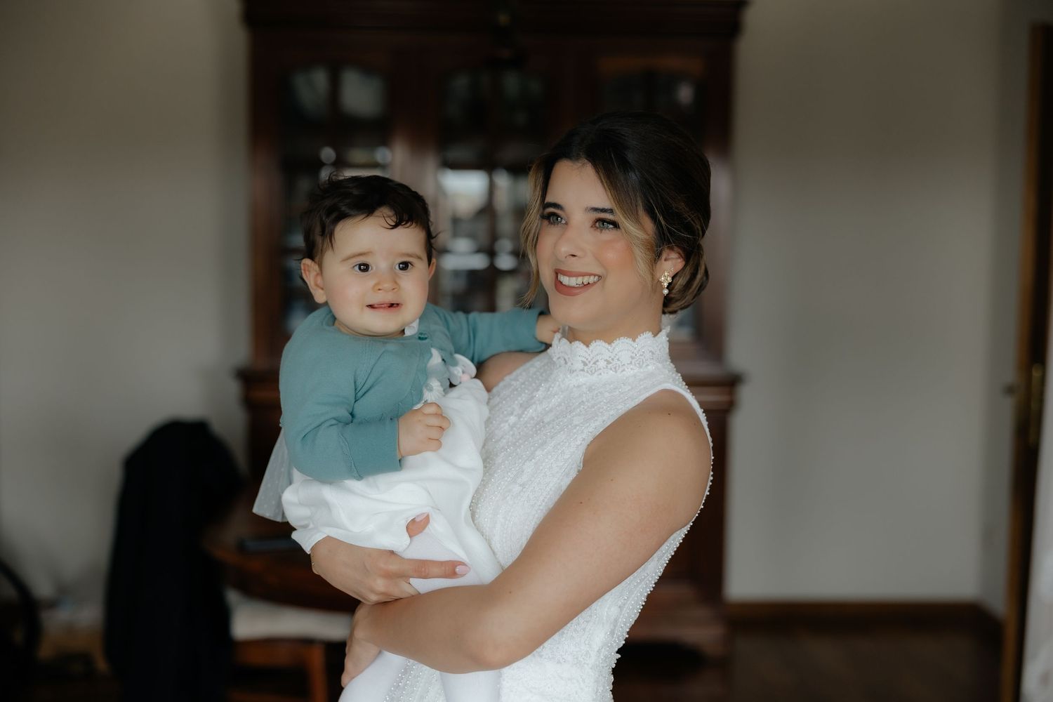Smiling bride in white beaded gown holds an adorable baby in a teal outfit before her wedding.