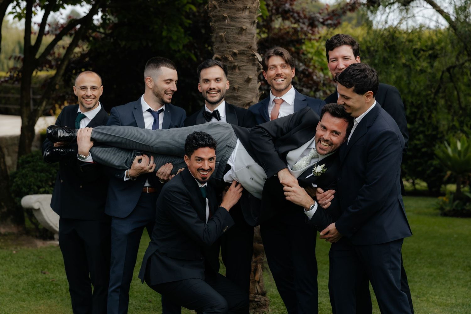 Groomsmen playfully lift groom horizontally in fun outdoor wedding party photo among palm trees.