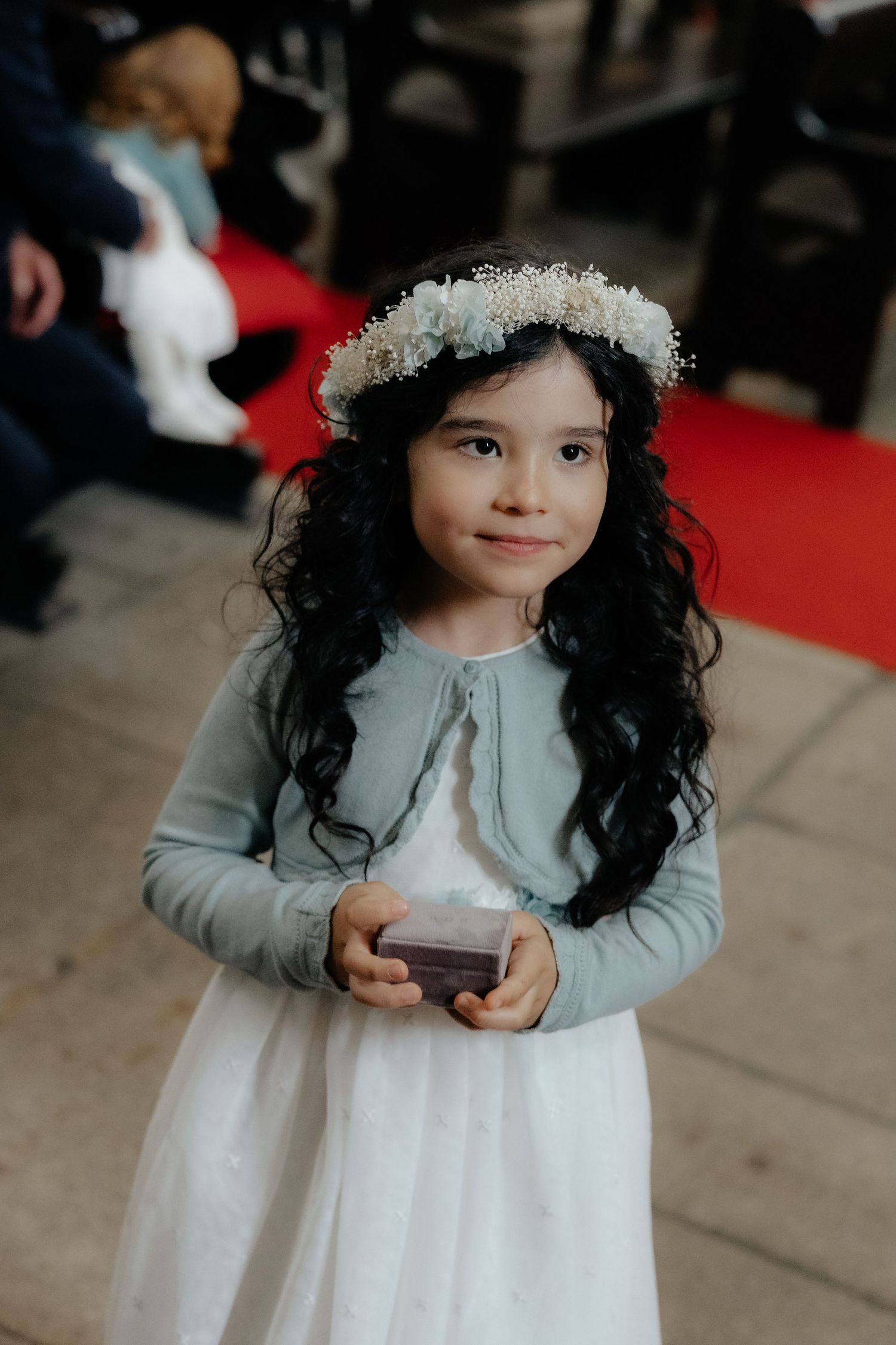 Young flower girl in white dress and floral crown holds a small box during a wedding ceremony.