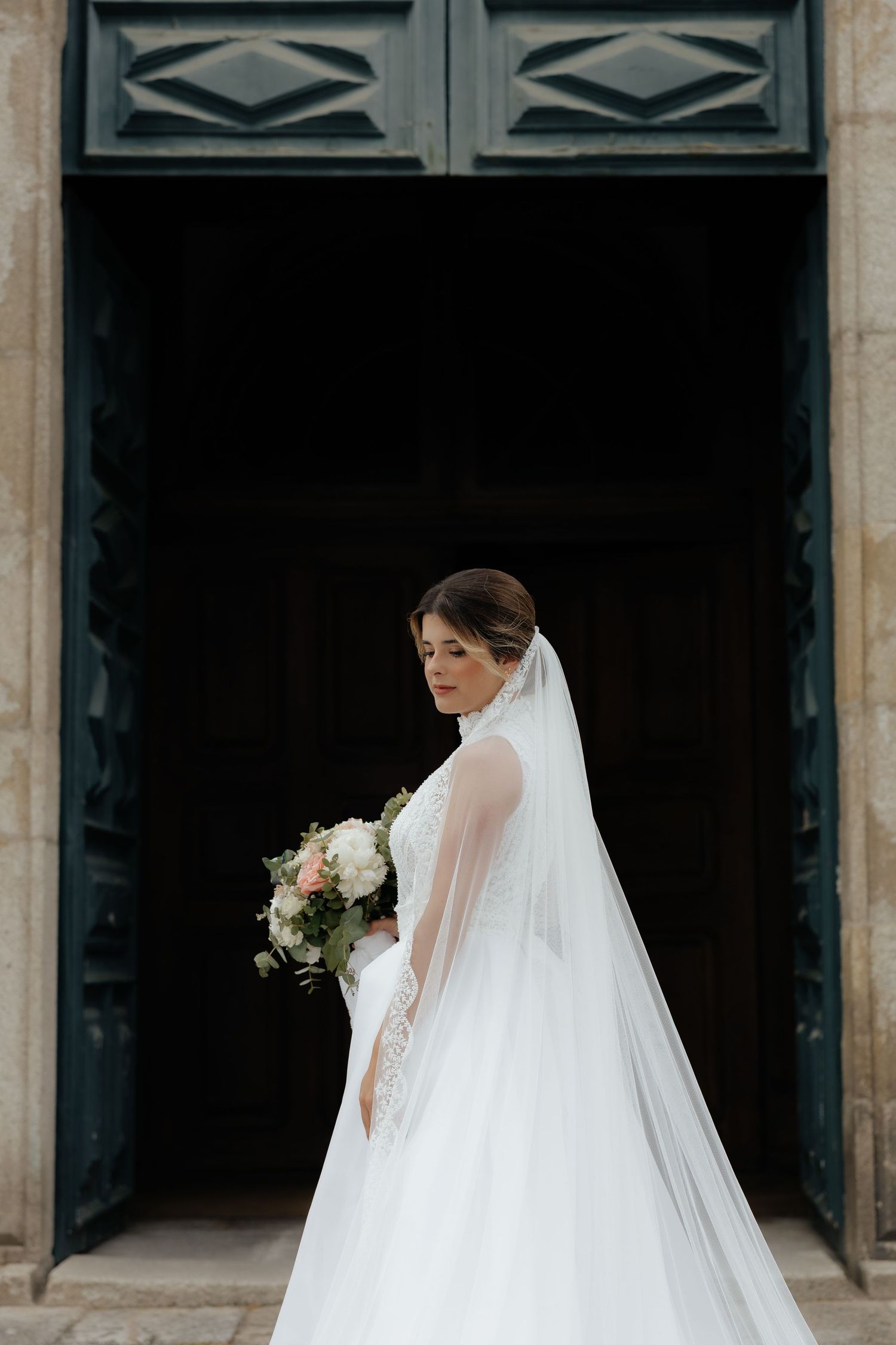 Bride in a full white gown and veil holding a bouquet while standing before large dark doors.