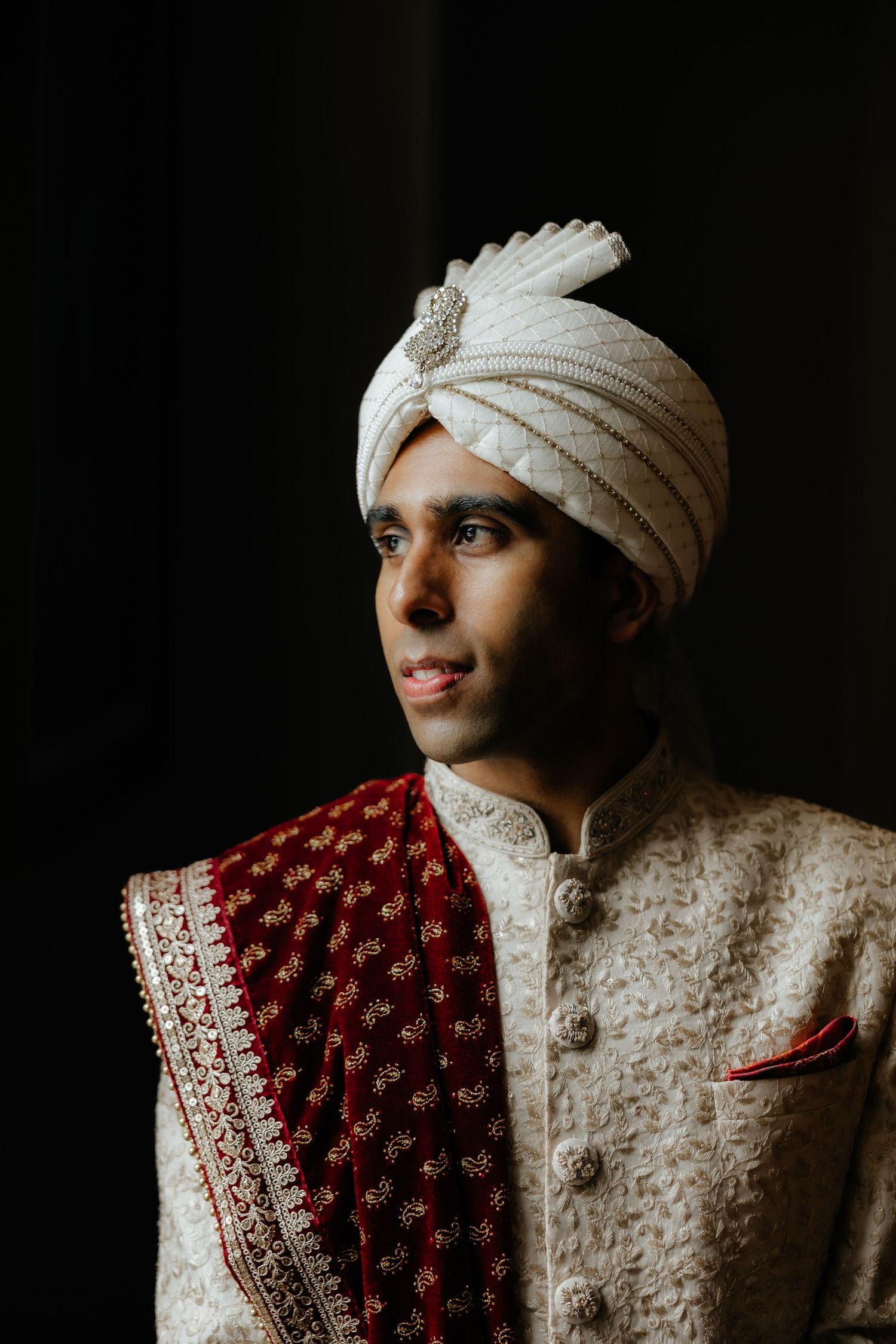 Indian groom in white sherwani and turban gazes thoughtfully during traditional wedding ceremony.