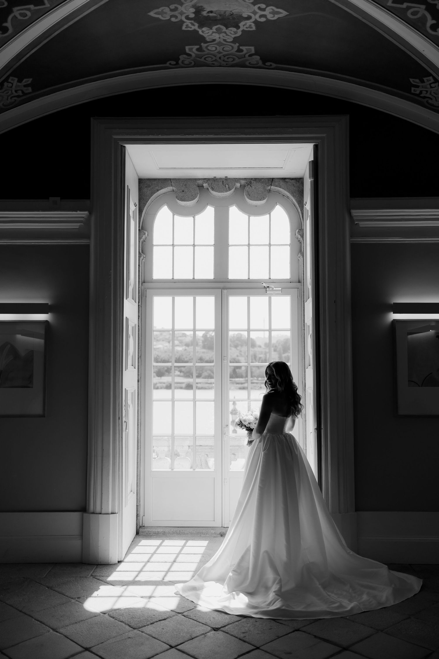 Dramatic black and white silhouette of bride holding bouquet before grand arched window in palace.