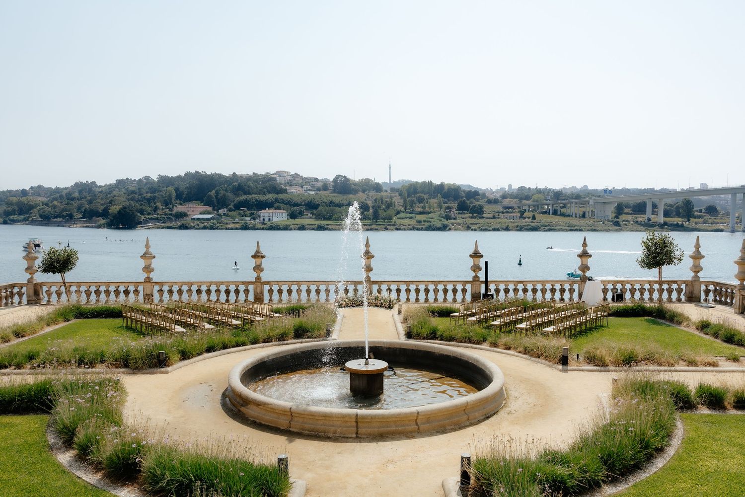 Elegant garden fountain with stone balustrade overlooking a serene river landscape on a sunny day.