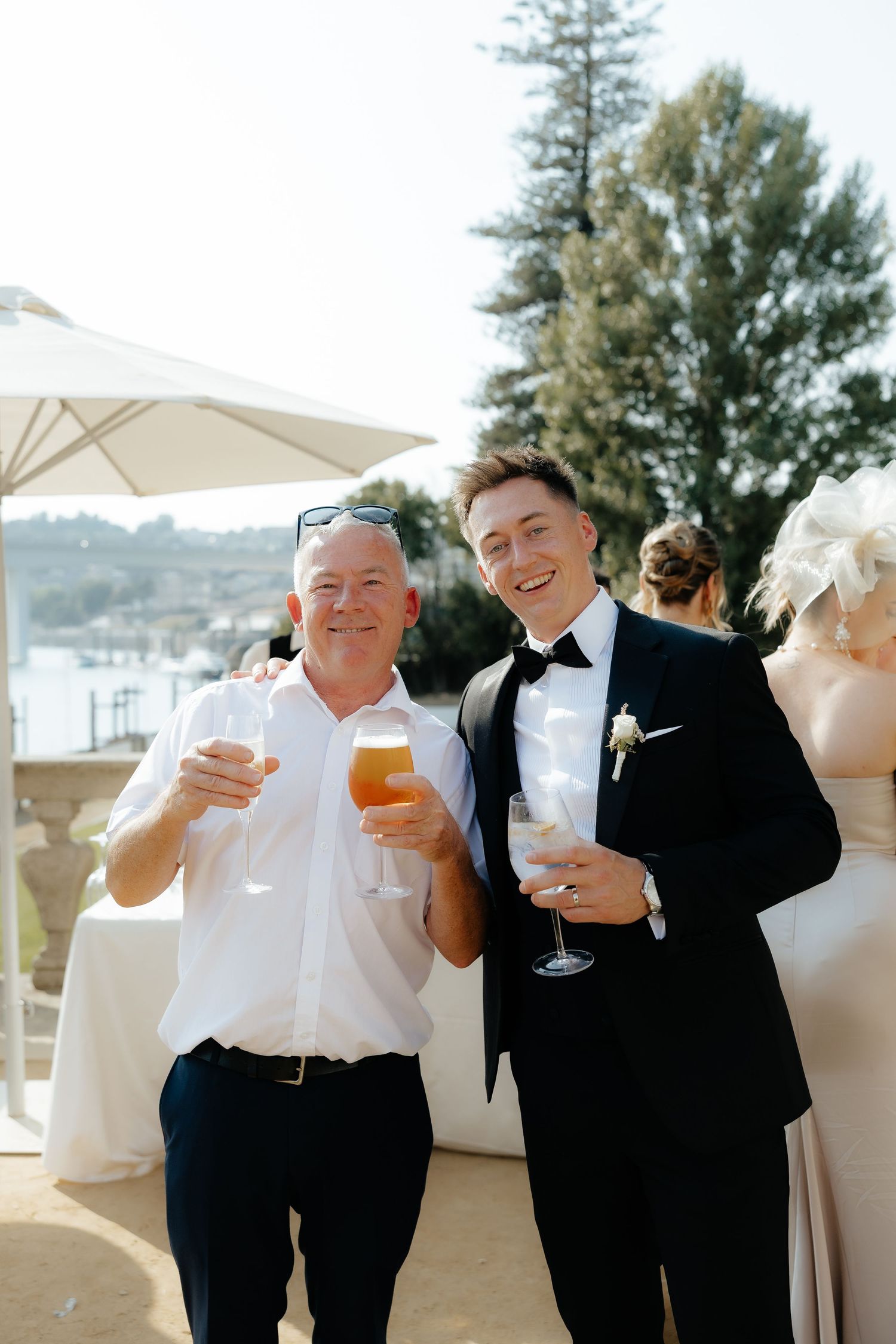 Two men smiling and raising glasses in a toast at an outdoor waterfront wedding reception.
