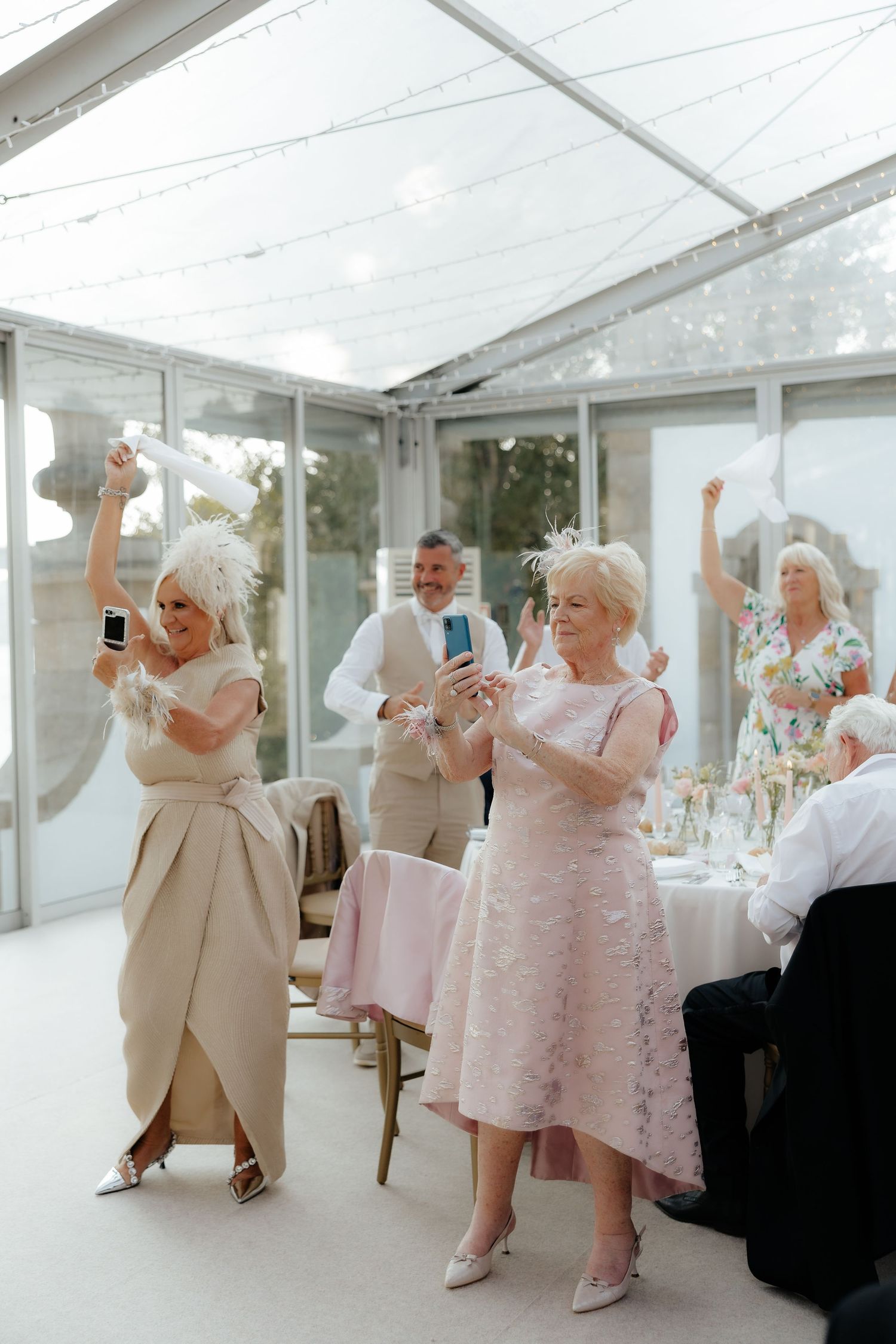 A group of wedding guests in pastel dresses and suits dance together in a bright white reception tent.